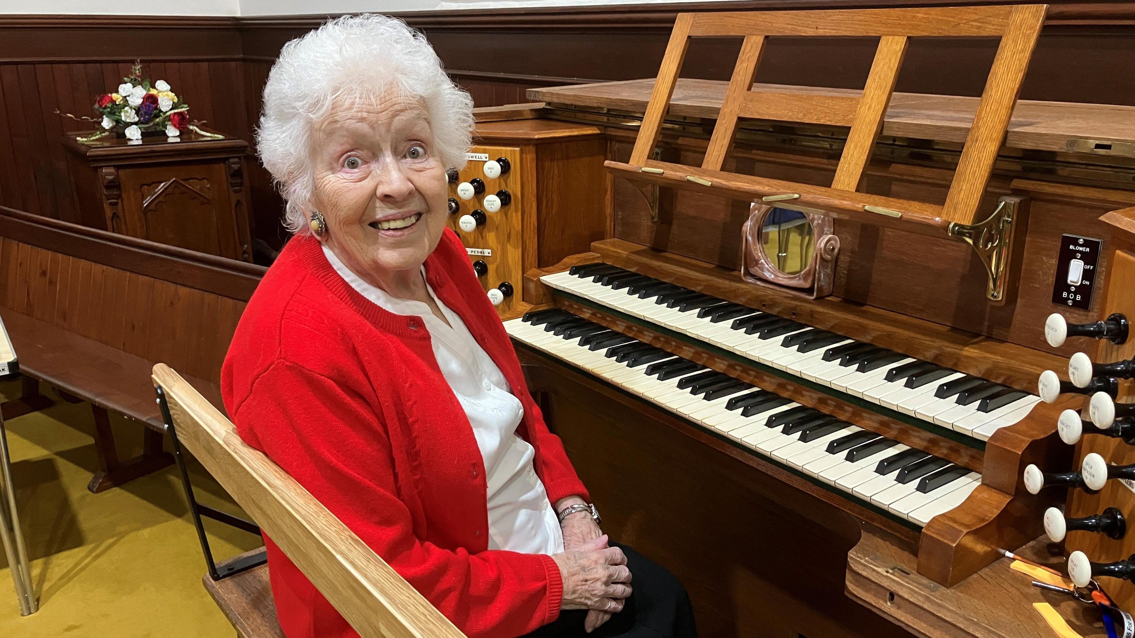 A grey-haired woman in a white blouse and red cardigan sits on a bench in front of a church organ