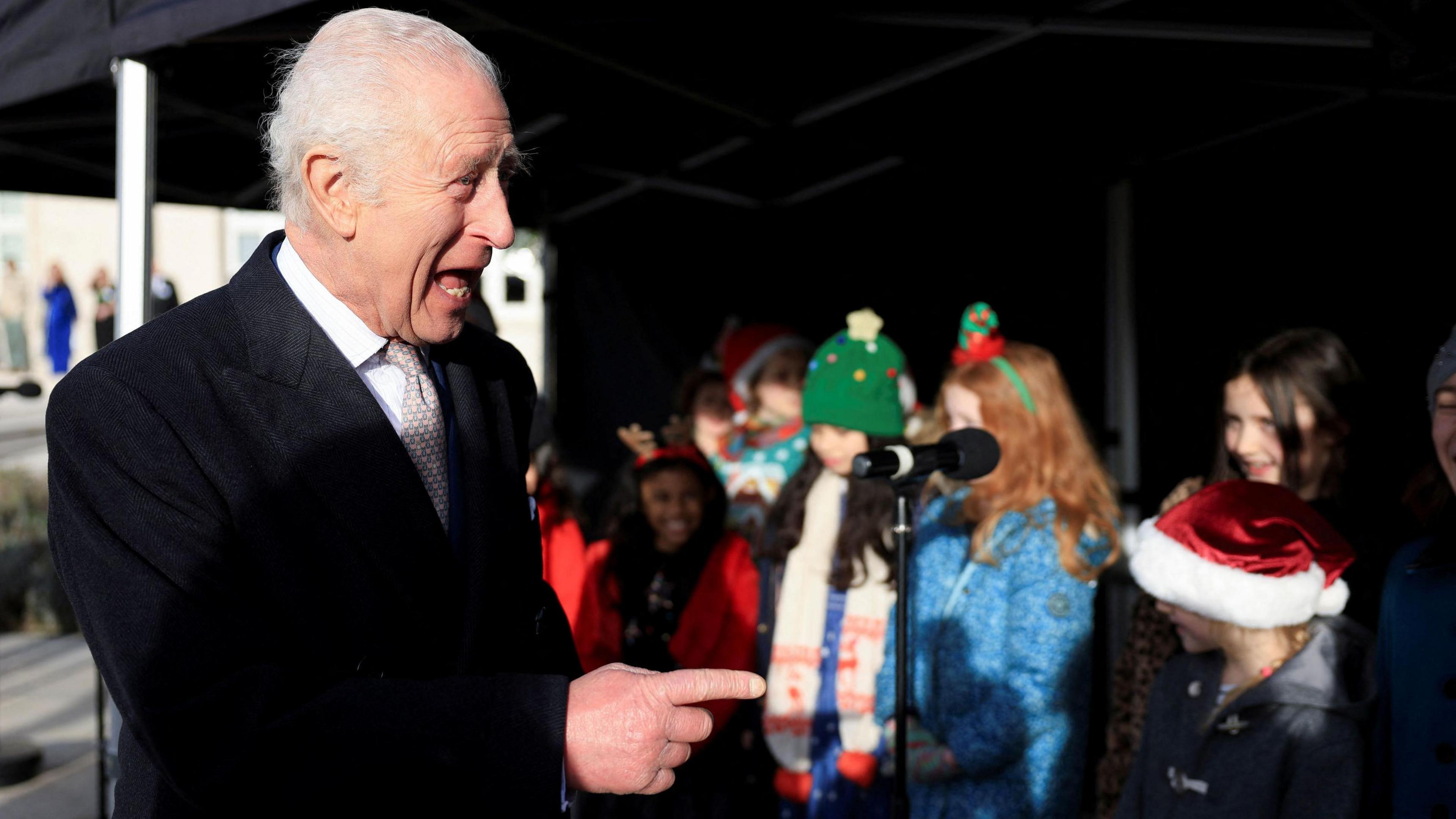 King Charles, wearing a black overcoat, points and laughs. In the background are a group of schoolchildren wearing festive hats. 