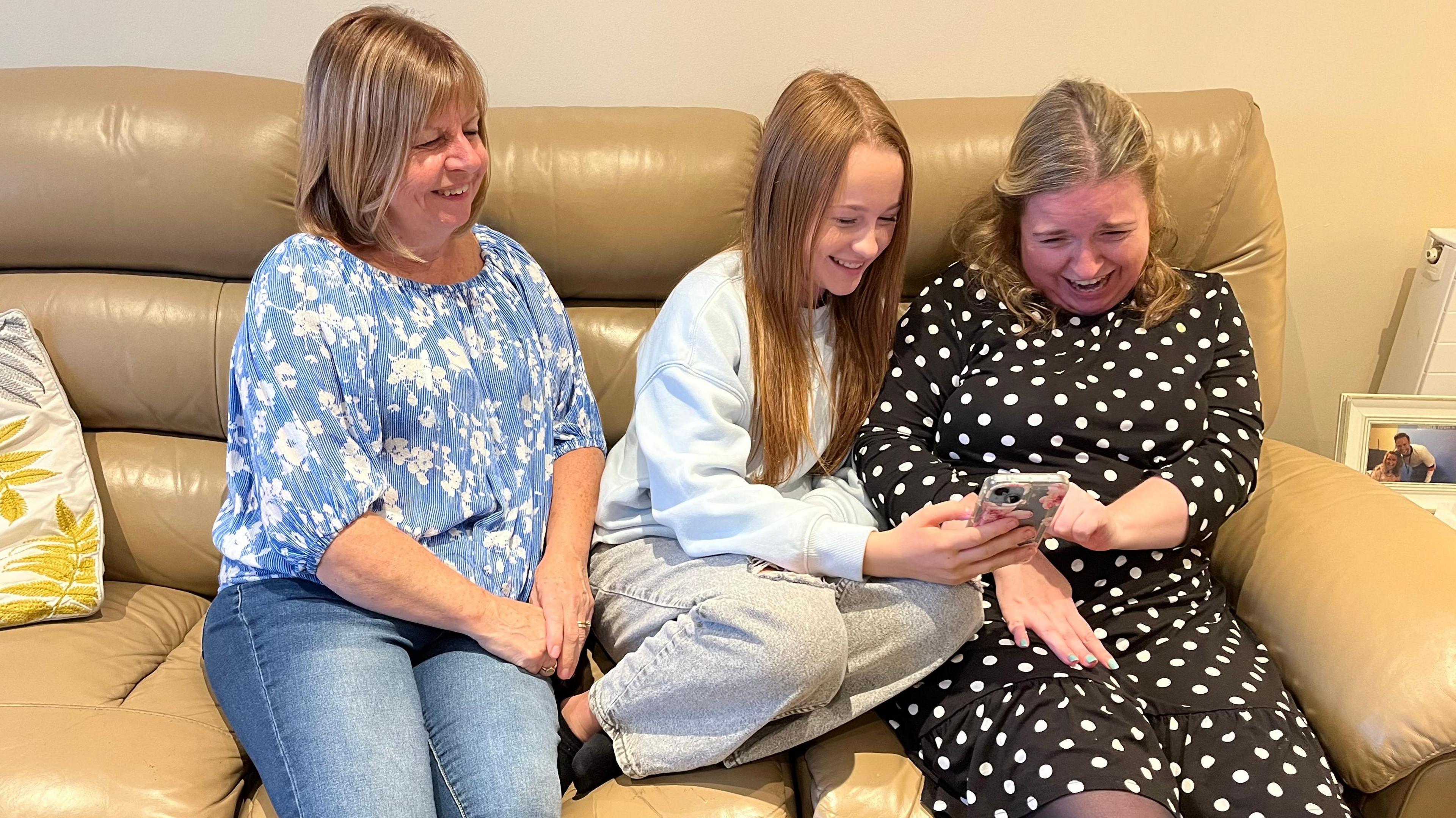 A woman of about 60, a teenage girl and a woman in her forties sit in a row on a tan leather sofa. The older woman wears a blue top with white flowers with a scoop neck and three quarter length sleeves, and pale blue jeans. She has a short blonde highlighted bob with a fringe and her hands are folded on her lap. She is smiling and looking towards a mobile phone which is being held by the teenager sitting in the middle. The teenager, who is also looking at the phone and smiling, has long dark blonde hair and is wearing a light blue crew neck sweatshirt and light grey jeans, her legs are tucked up onto the sofa. The woman to her right has shoulder-length hair, slightly curled and pulled back from her face. She is touching the phone with a finger of her left hand and looking at the screen while laughing. Her other hand lies in her lap. She is wearing a black dress with long sleeves and white polka dots. The phone case is grey with pink flowers. To the right of the picture, a framed picture of two people is visible, and the corner of a radiator. To the left, part of a cream and mustard patterened cushion can be seen on the sofa.