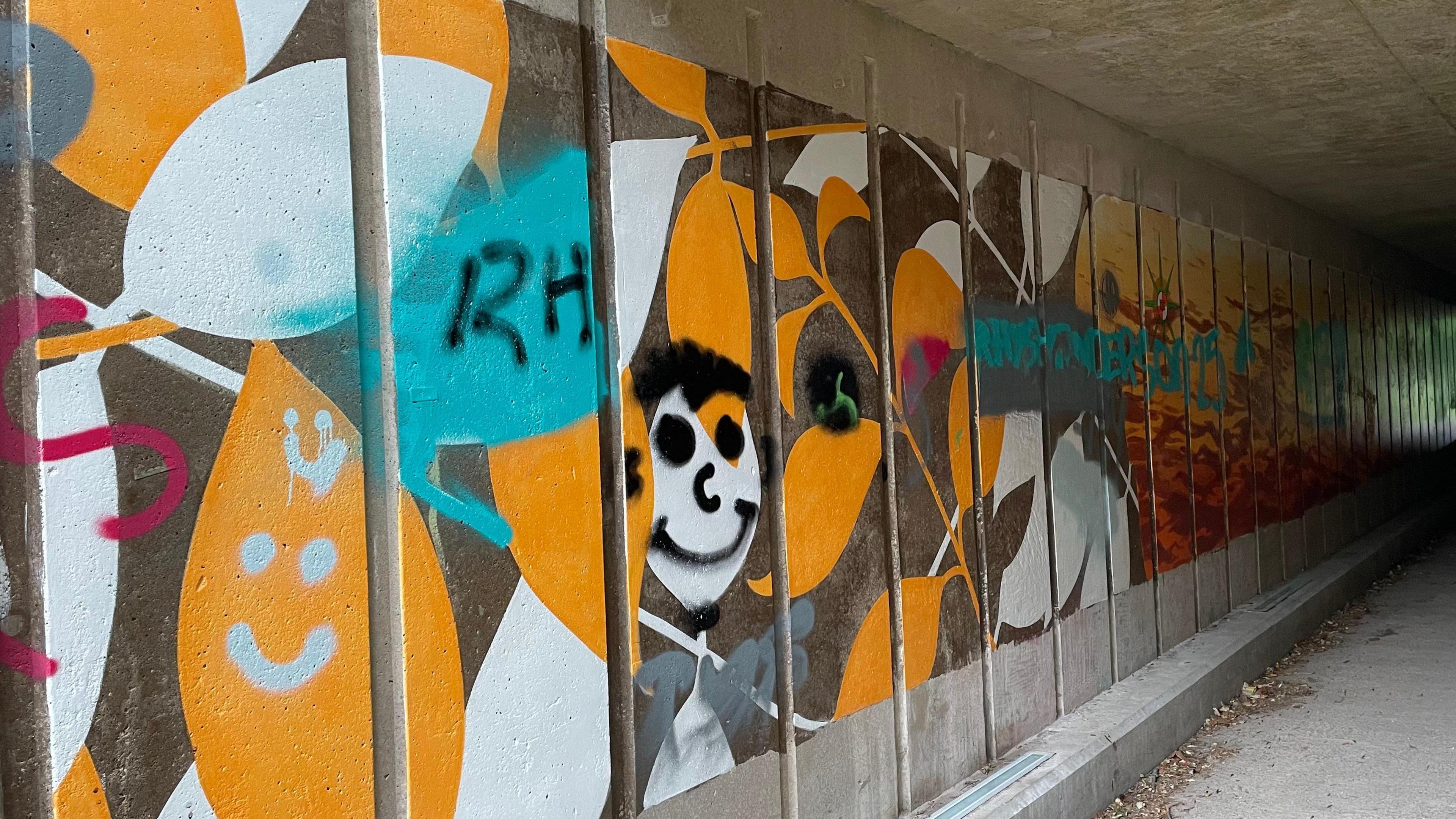 A mural along a wall in a tunnel. It has been defaced with different colours of graffiti.