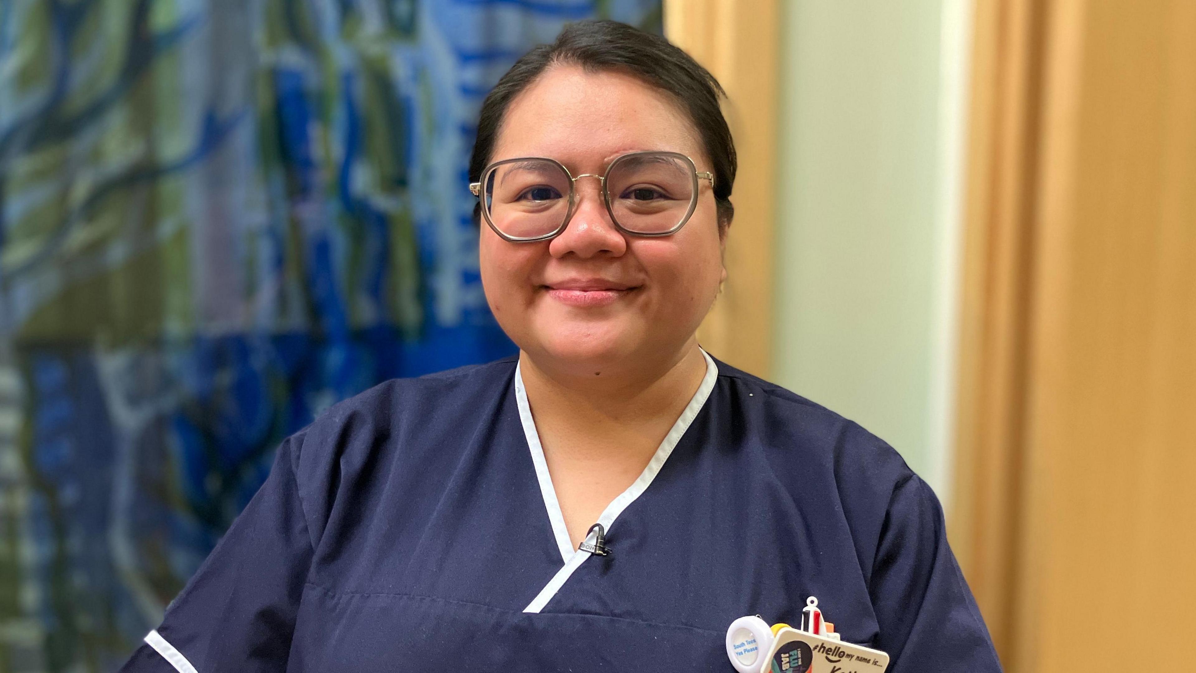 Katha Deguzman is wearing a navy blue nurses tunic and smiling at the camera. She wears round, metal rimmed glasses and her dark hair is tied back in a neat bun. She is from the Philippines. 