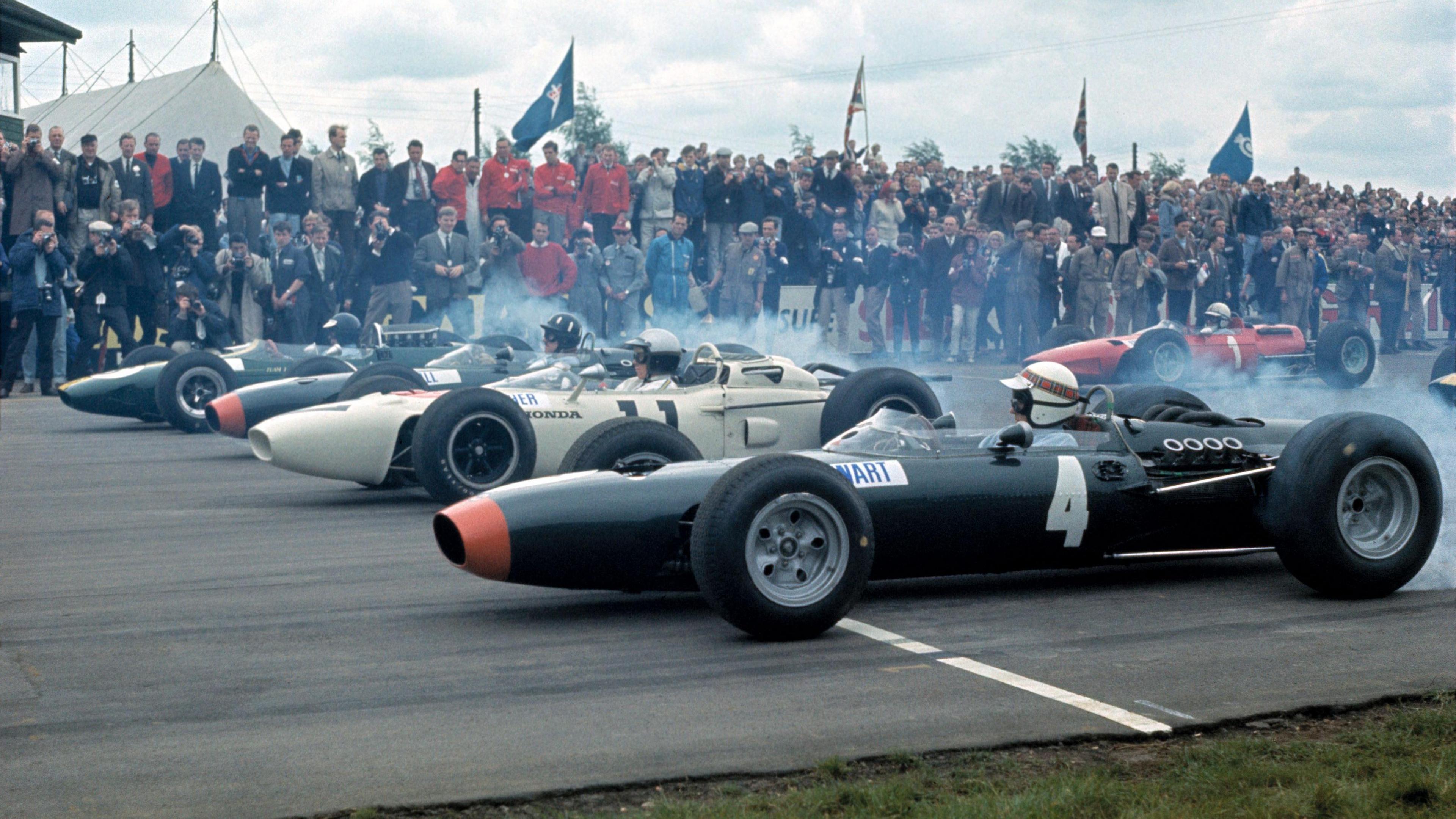 Tyre smoke fills the air as the field moves off for the 1965 race at Silverstone with Jackie Stewart's BRM nearest the camera, then Richie Ginther's Honda, Graham Hill's BRM and Jim Clark's Lotus, with the Ferrari of John Surtees in the second row