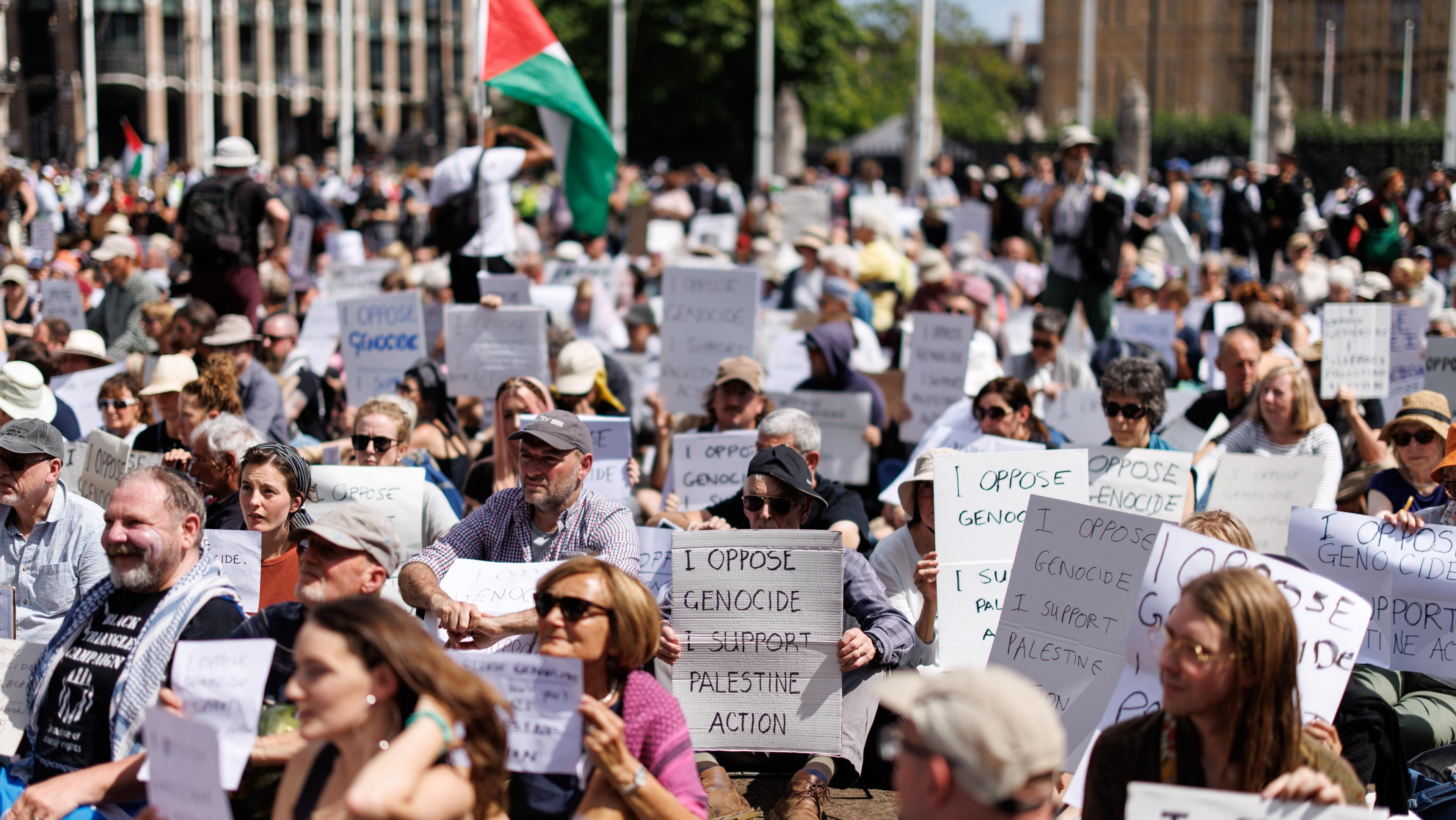 A large group of protesters are sitting down, many are holding up pieces of card or paper reading 'I oppose genocide I support Palestine Action'