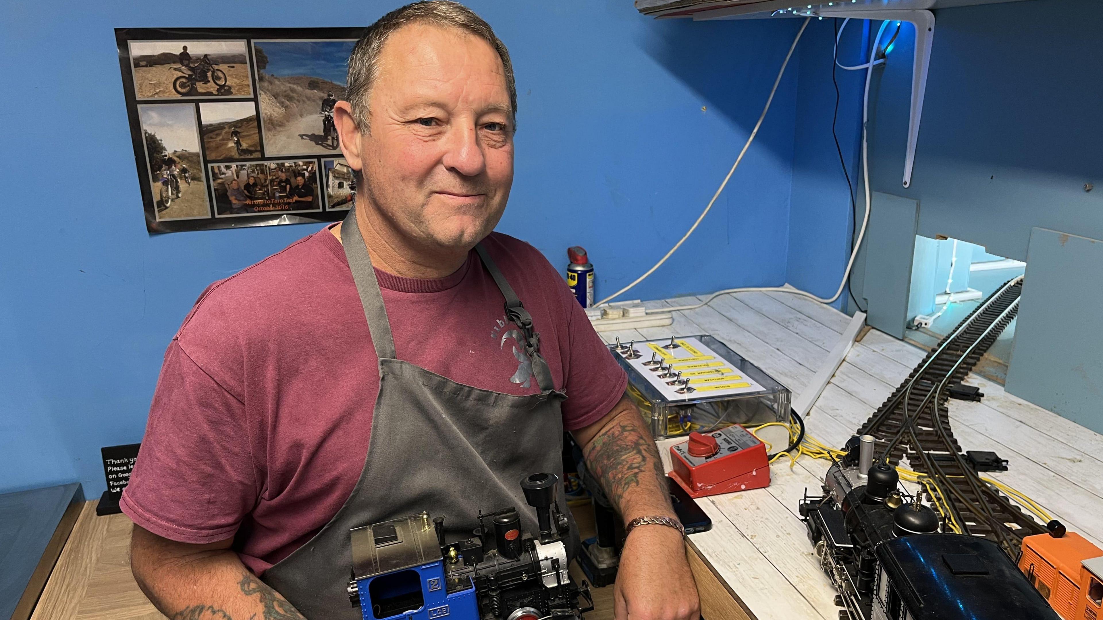 Dave Wheeler wearing an apron and holding a model train engine with track in the background