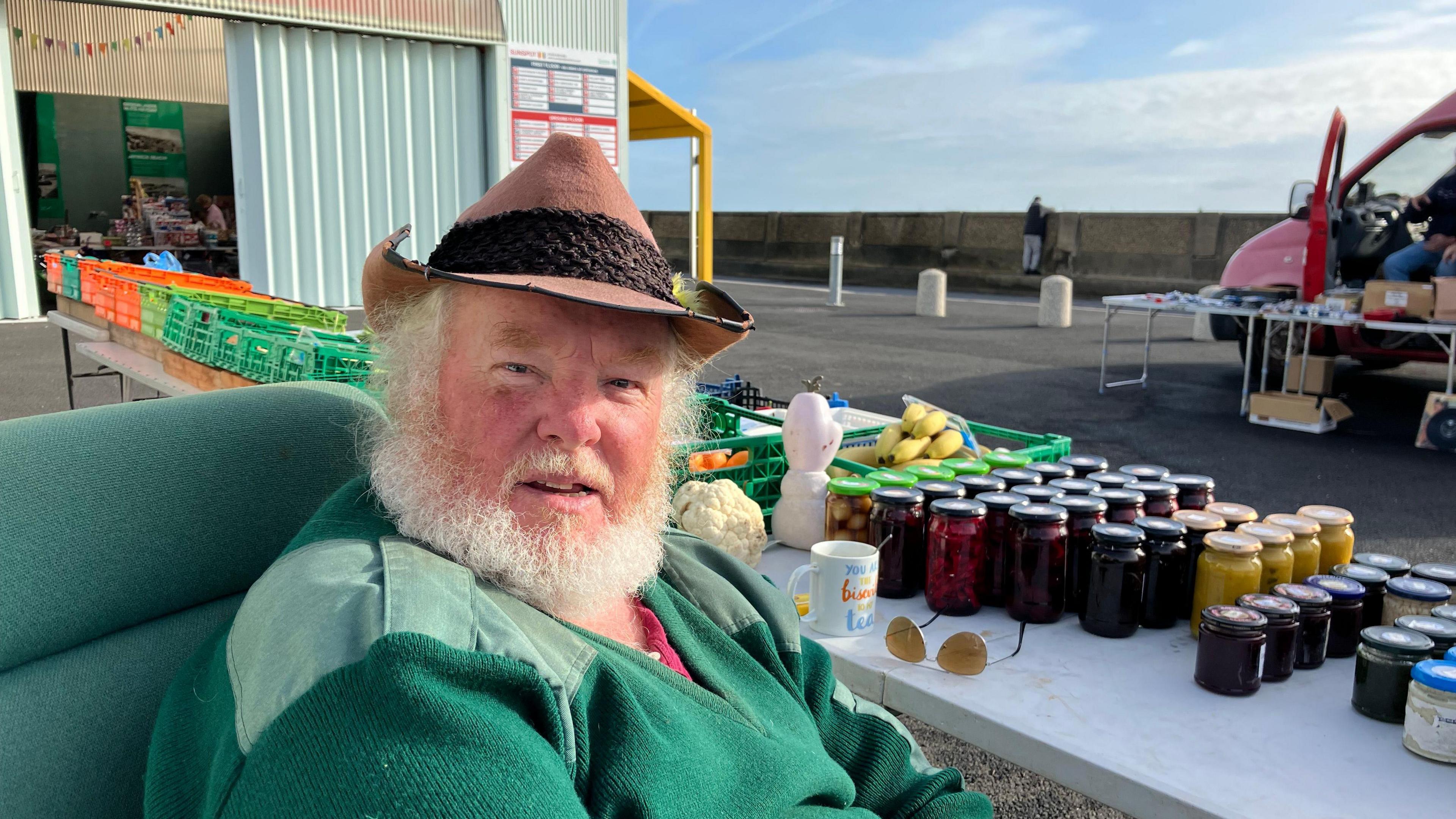 A man with a brown hat and a green jumper sitting in front of some jars and fruit & vegetables on a table