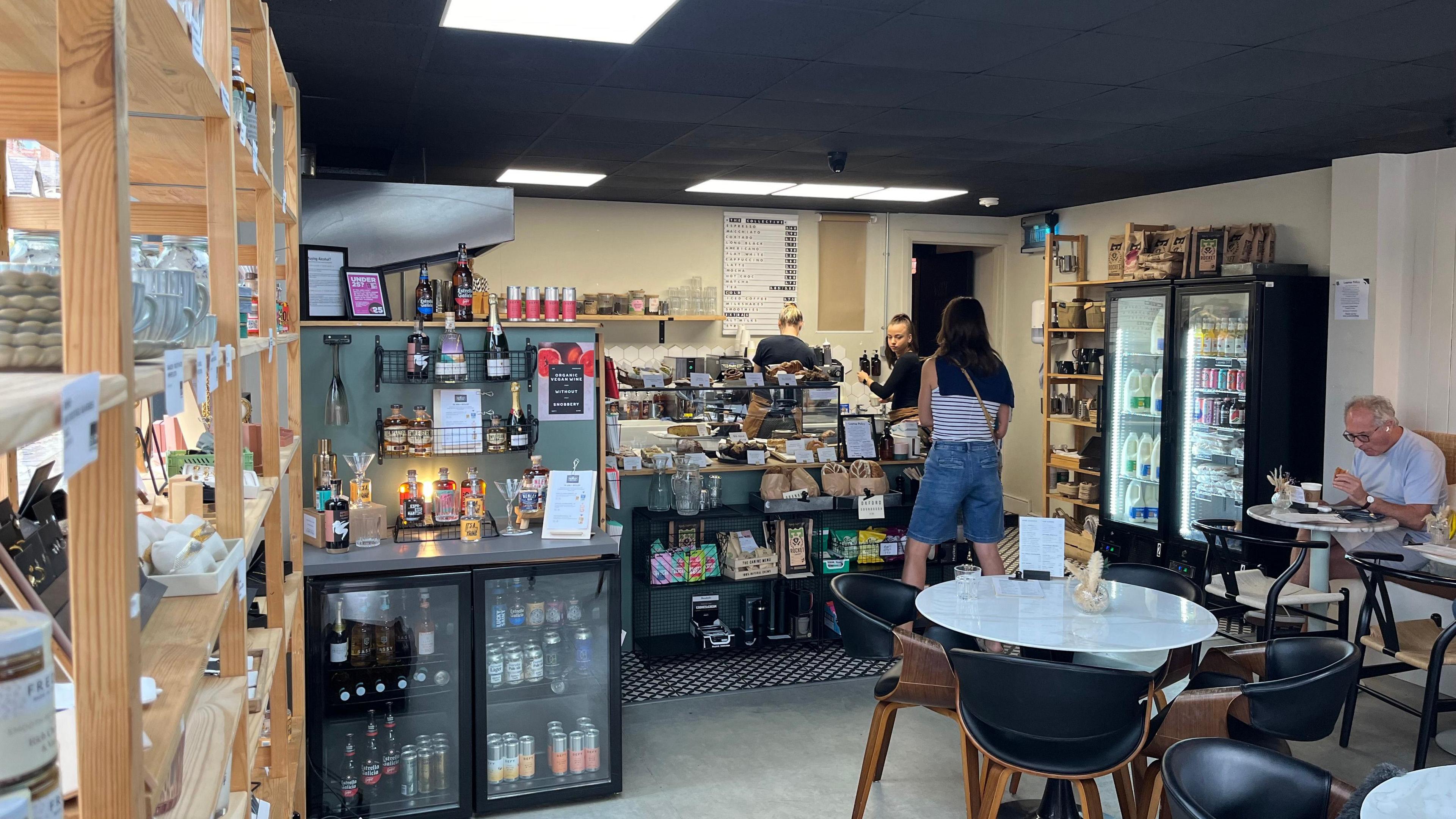 A general view of the coffee shop, there are some tables with chairs around the, and a counter at the front with cakes on display. It is a reasonably small area.