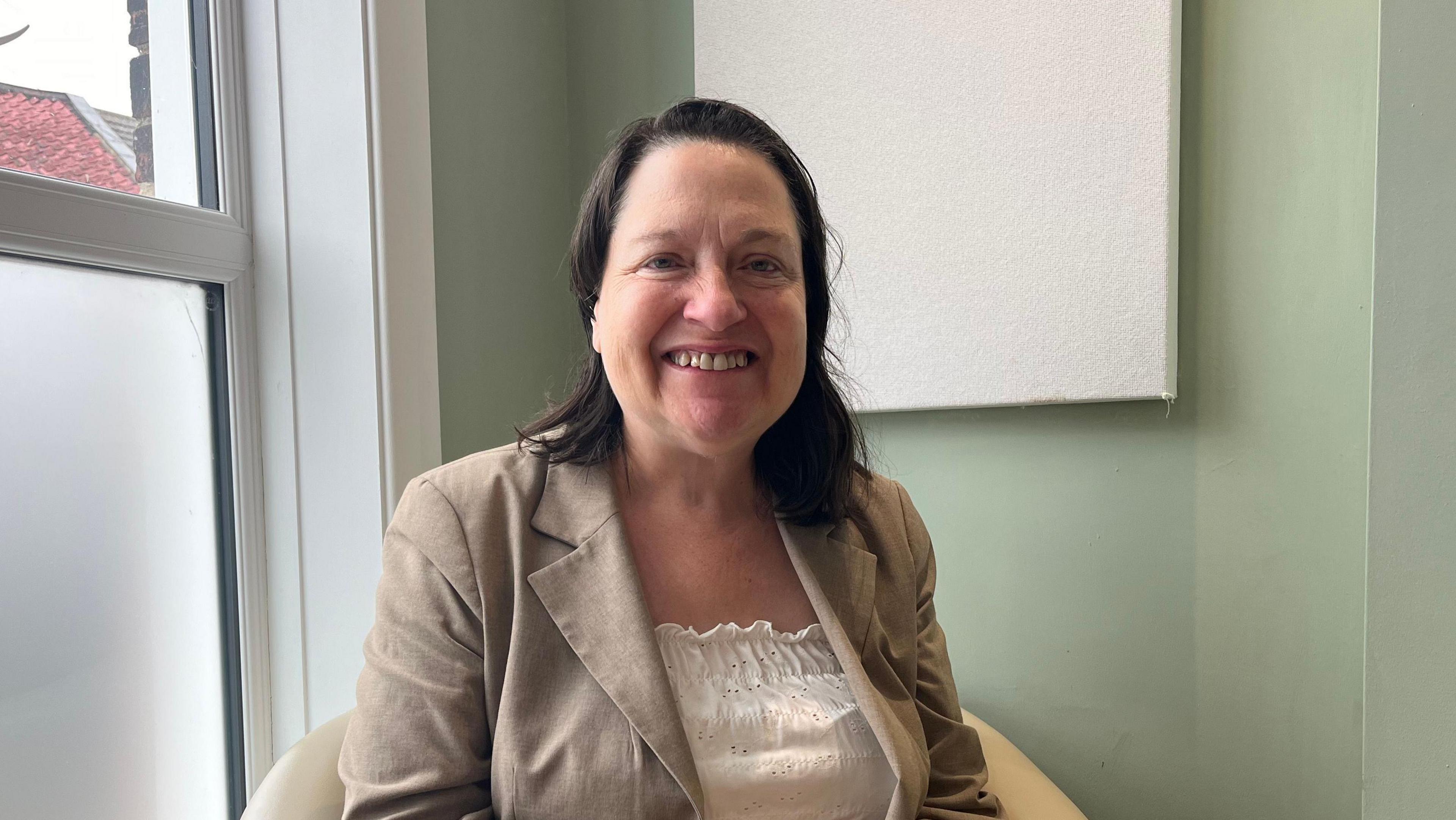 Alexandra Kemp smiles at the camera as she sits in a cream coloured chair. She is wearing a cream jacket and top. Her hair is worn in a dark brown bob. 