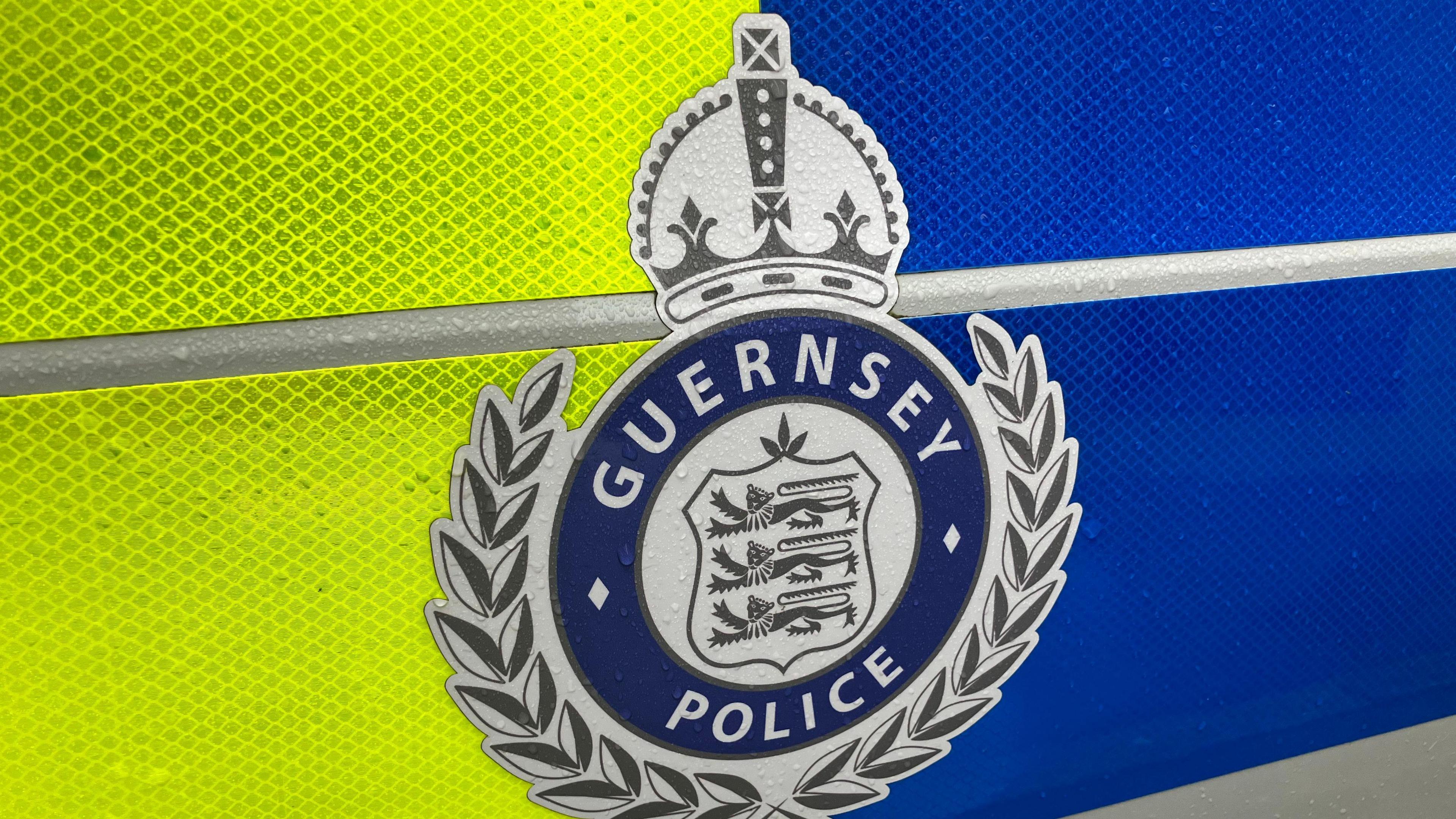 The side of a yellow and blue police vehicle. At the centre is a Guernsey Police badge which is silver and dark blue.