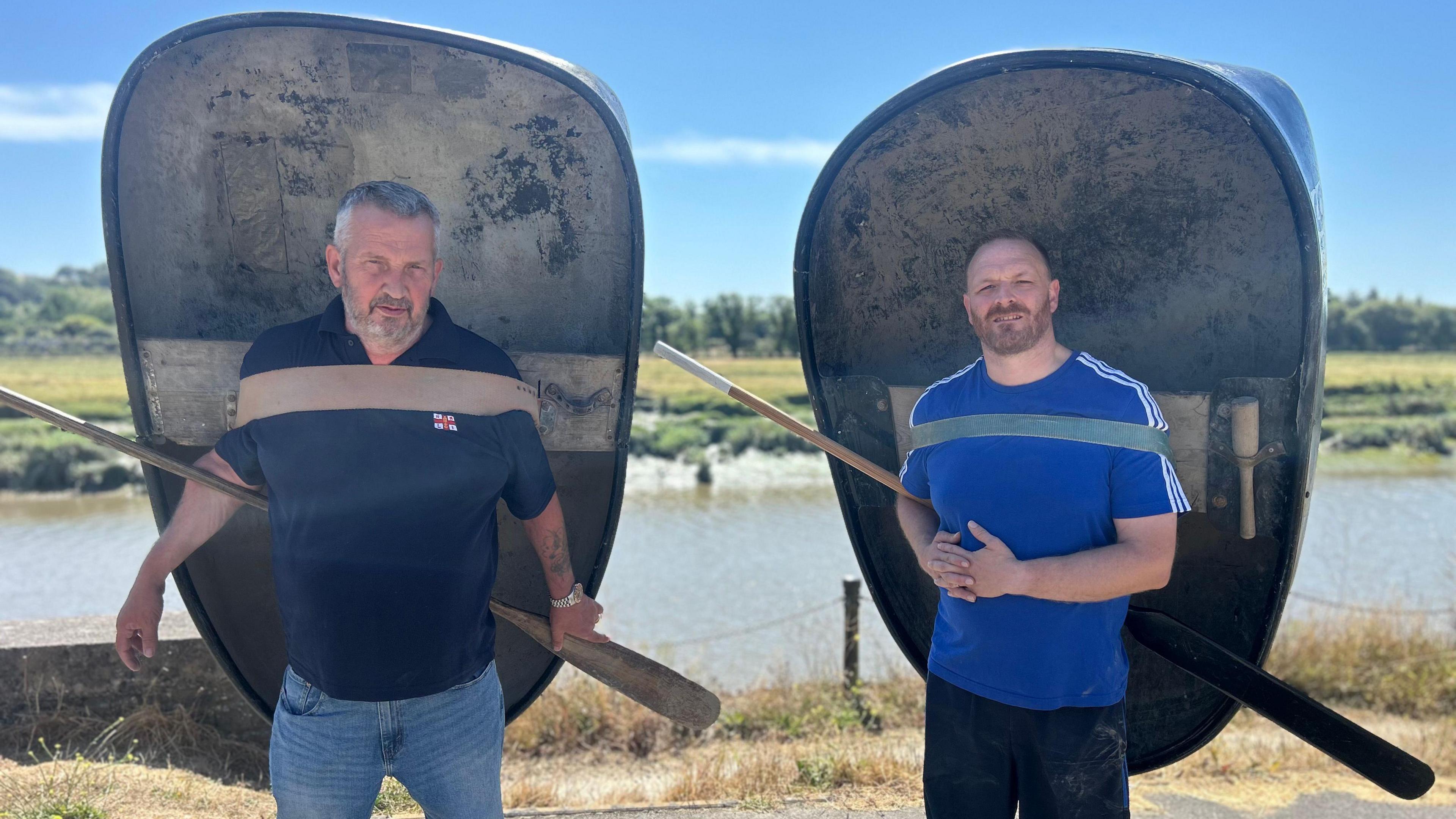 Andrew on the left wearing a navy RNLI polo shirt and blue jeans. Keith is on the right wearing a blur t-shit and navy trousers. Both have coracles; small rounded boats attached to their backs and holding oars. The river Tywi is behind them.
