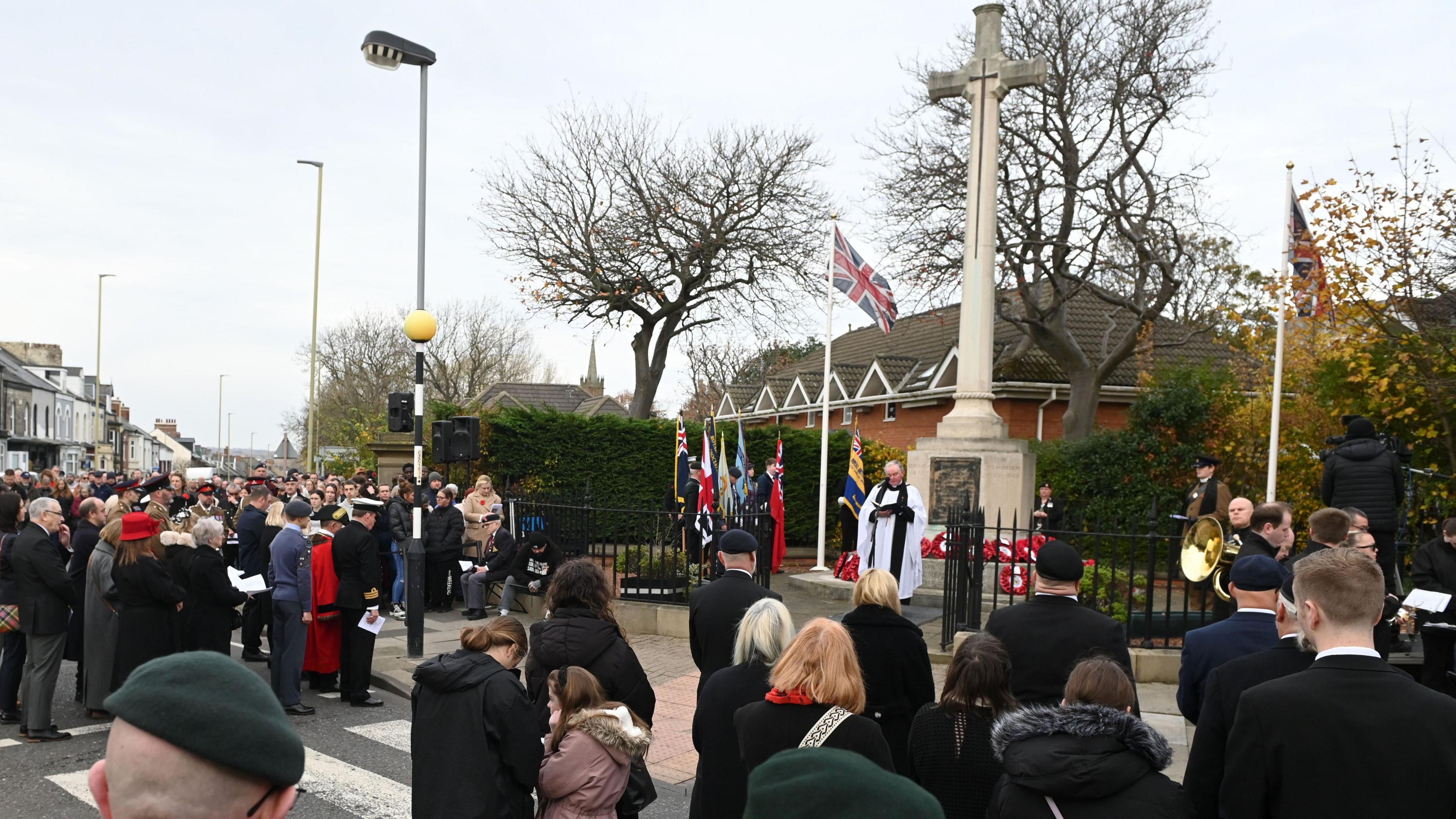 A priest wearing white robes stands by the war memorial in Westoe and holds a Remembrance Service. The streets are lined with members of the public, veterans and armed forces personnel.