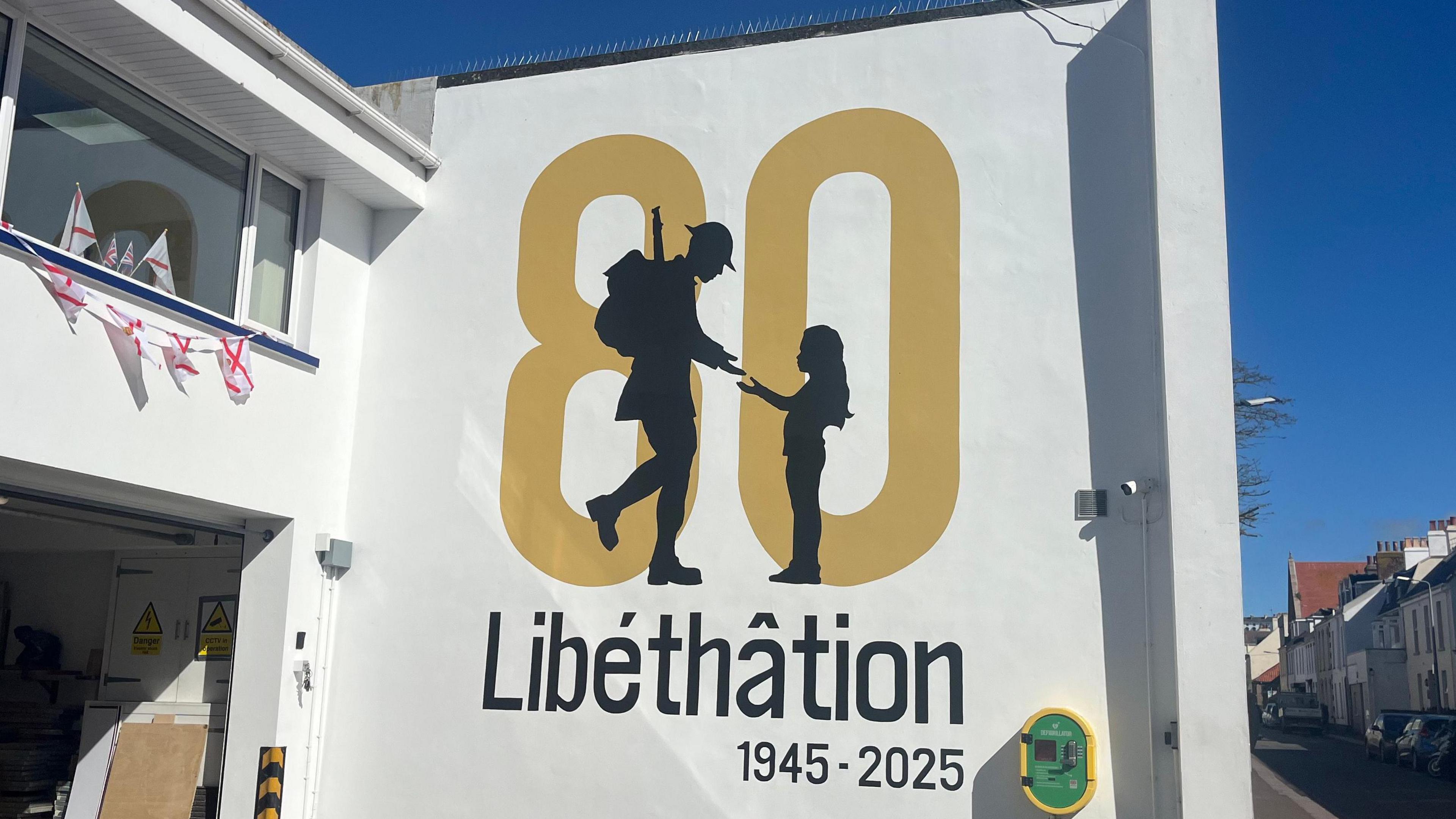 The mural shows a giant number 80 in gold-yellow lettering, with a silhouette of a soldier and a child. The soldier is reaching for the child's hand. It reads Libethation and below it, 1945 to 2025.