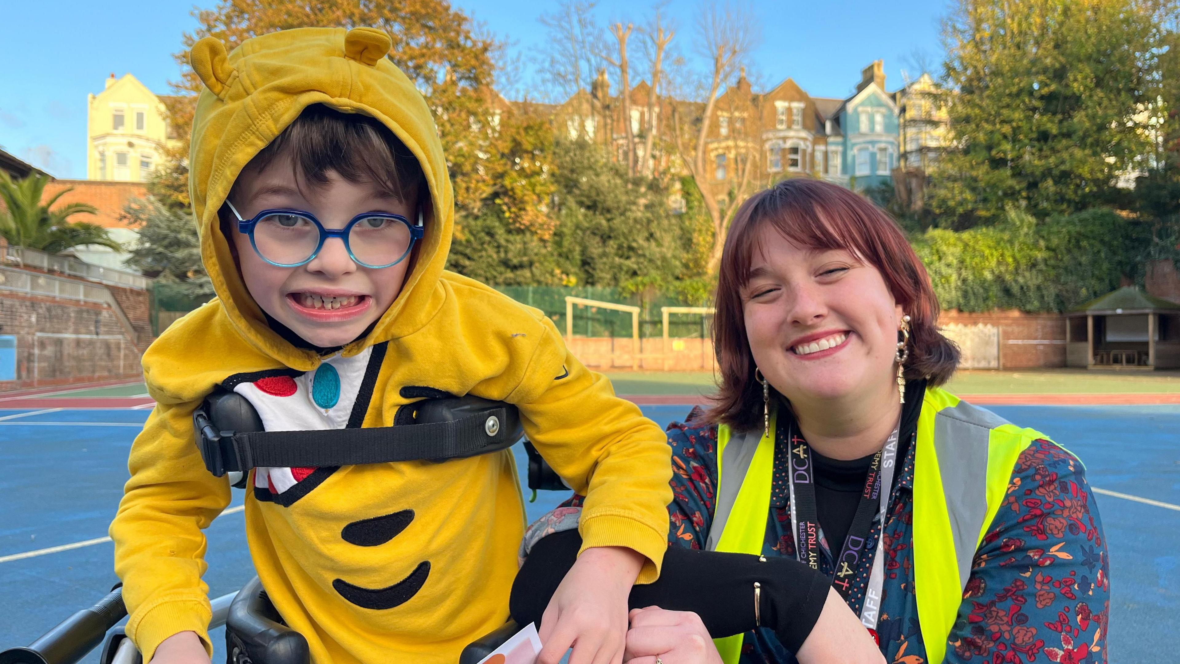 A boy with a harness to help him stand wearing glasses and a Pudsey hoodie standing next to a teacher with shoulder-length red hair and a high vis jacket