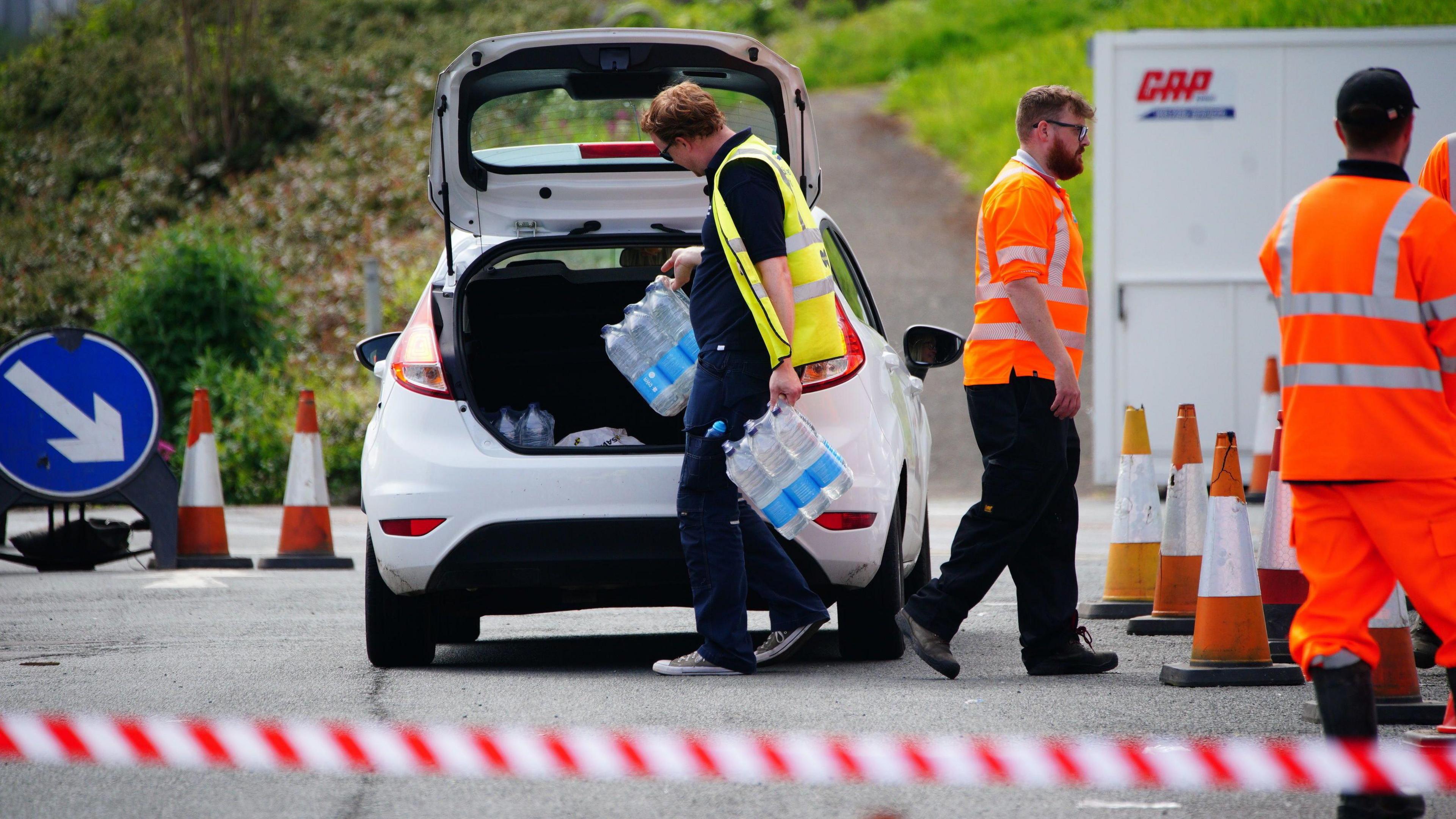 A man in a high-viz coat loads packs of bottled water into the boot of a white car.  