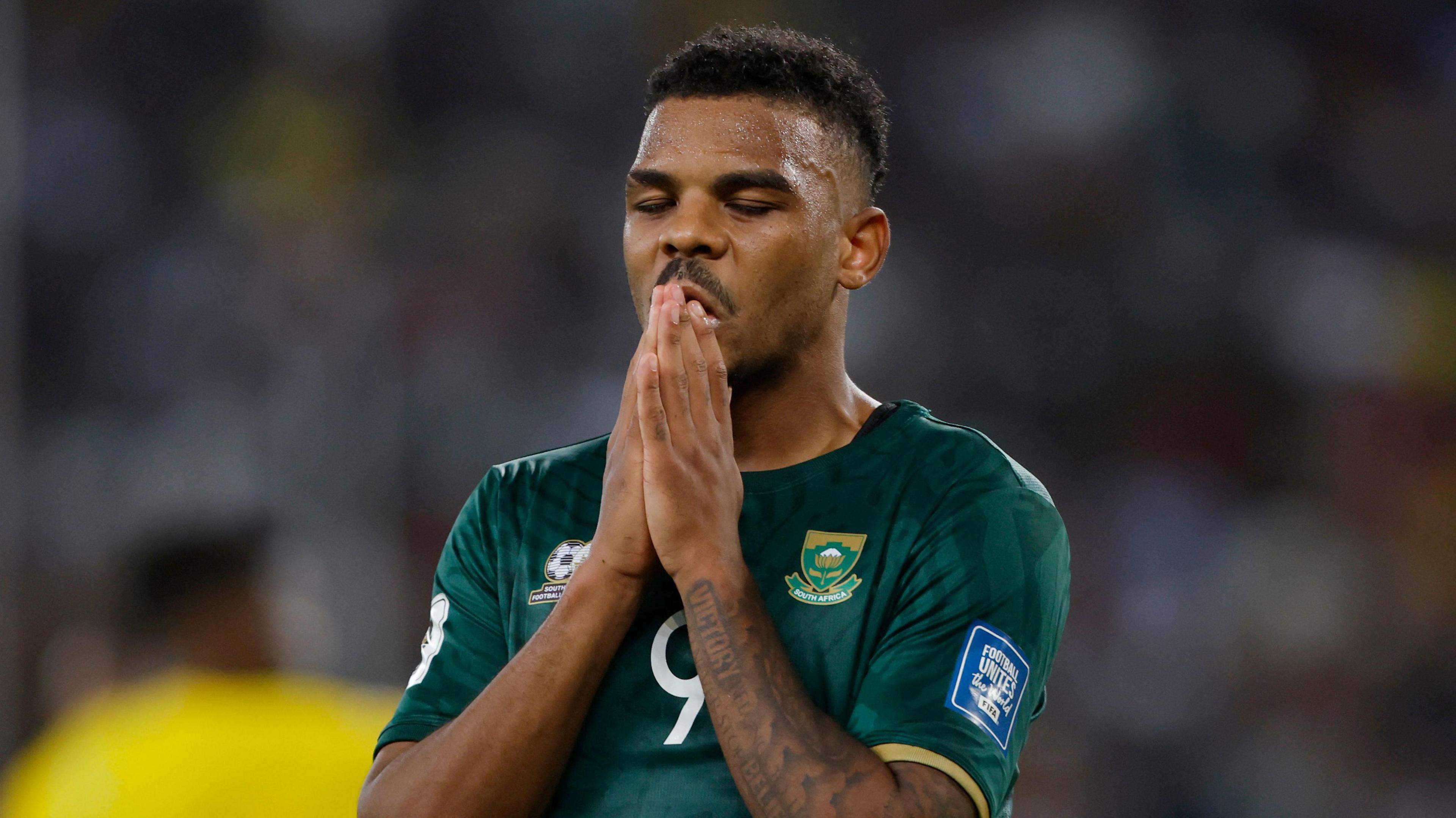 South Africa forward Lyle Foster, seen from stomach up wearing a green South Africa shirt, closes his eyes, puts his hands together in a prayer position and raises his hands to his lips during a football match