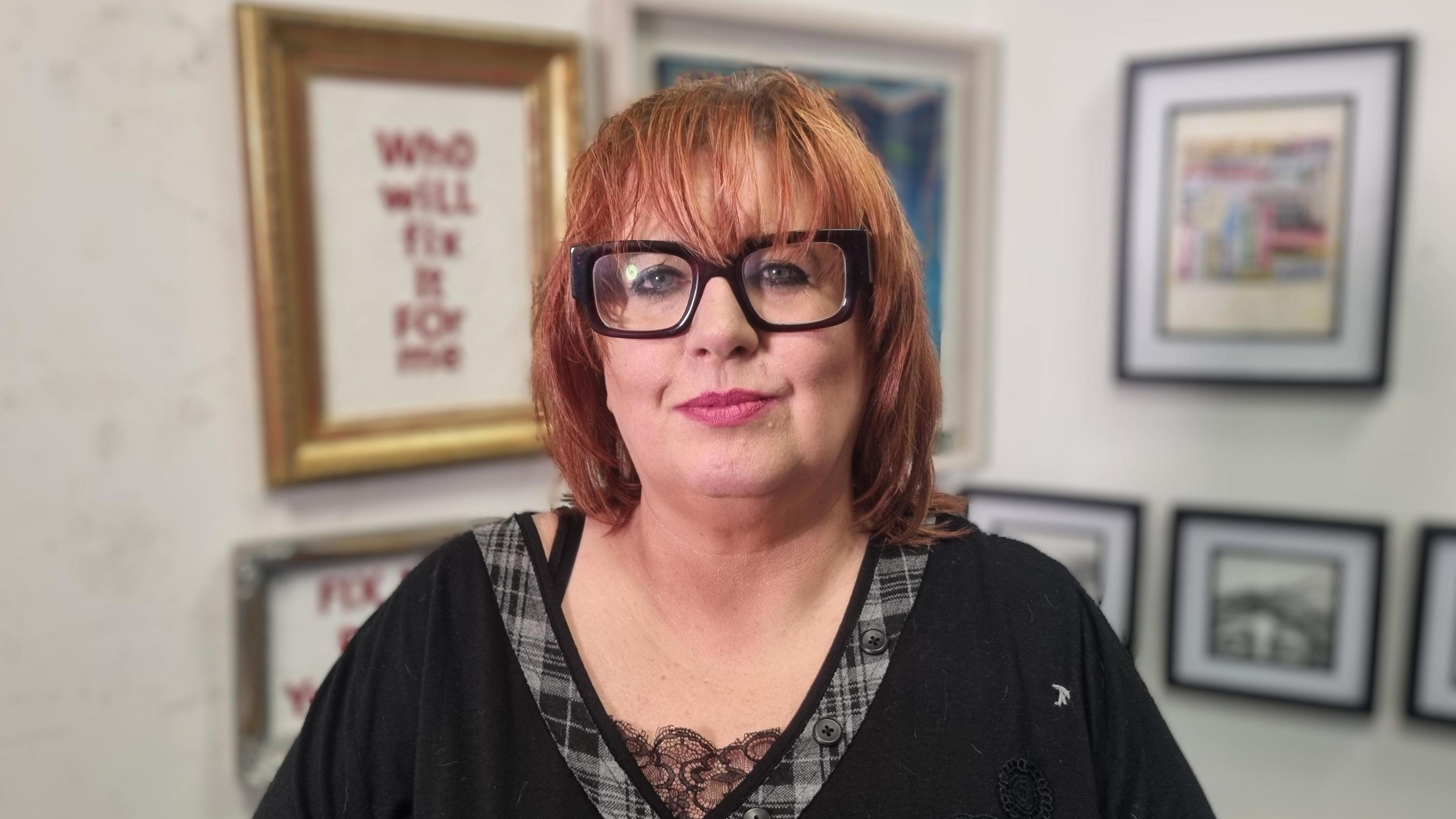 Artist Lesley Cherry, who has auburn hair and is wearing glasses, pictured standing in front of some of her artwork hanging in frames on a white wall in her studio in Belfast city centre