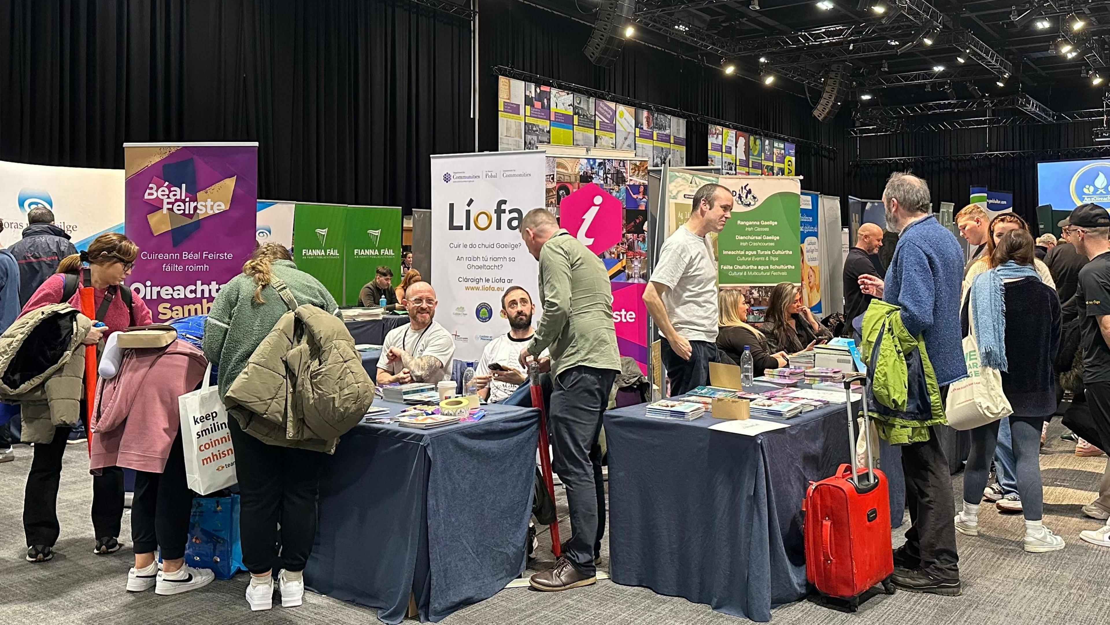 Several stalls set up at the event with large banners and tables covered with a blue tablecloth, and various posters on the tables. There are people sitting behind the stalls as well as people looking at the products on the table.