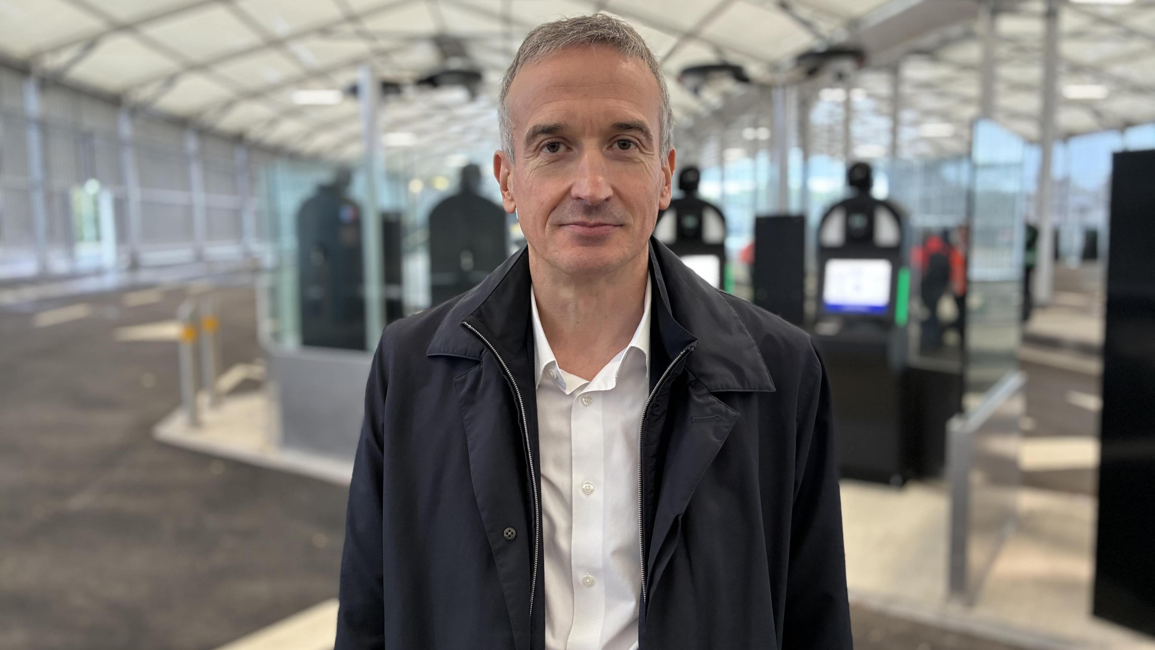 Close up of Eurotunnel CEO Yann Leriche standing in a hanger in Folkestone with biometric machines behind him. He has short grey hair and is wearing a black jacket and white shirt.