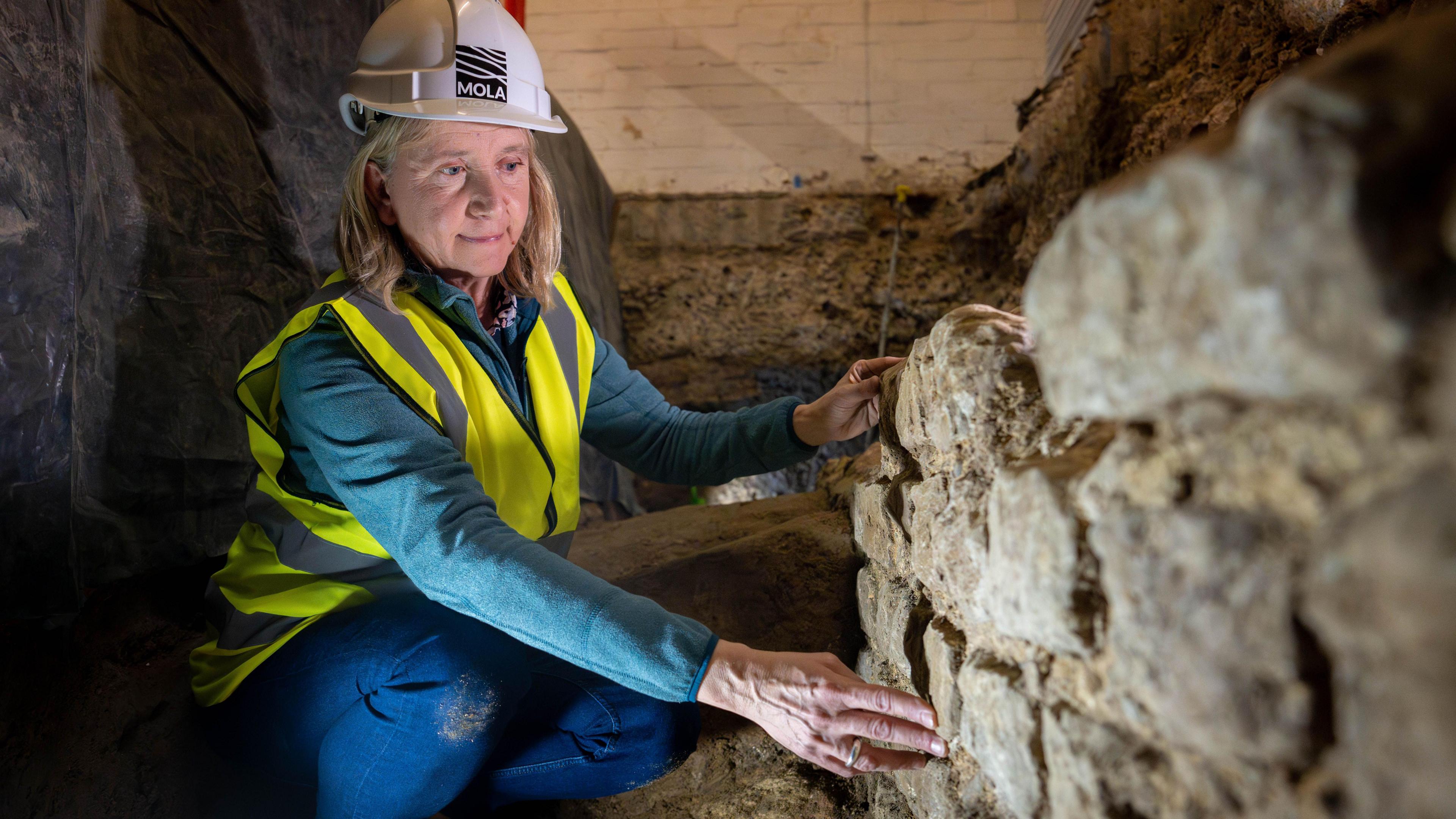 Archaeologist Sophie Jackson in a yellow high vis vest and white hard hat crouching next to a large piece of Roman wall about one metre hight made up of several layers of large grey stones. 