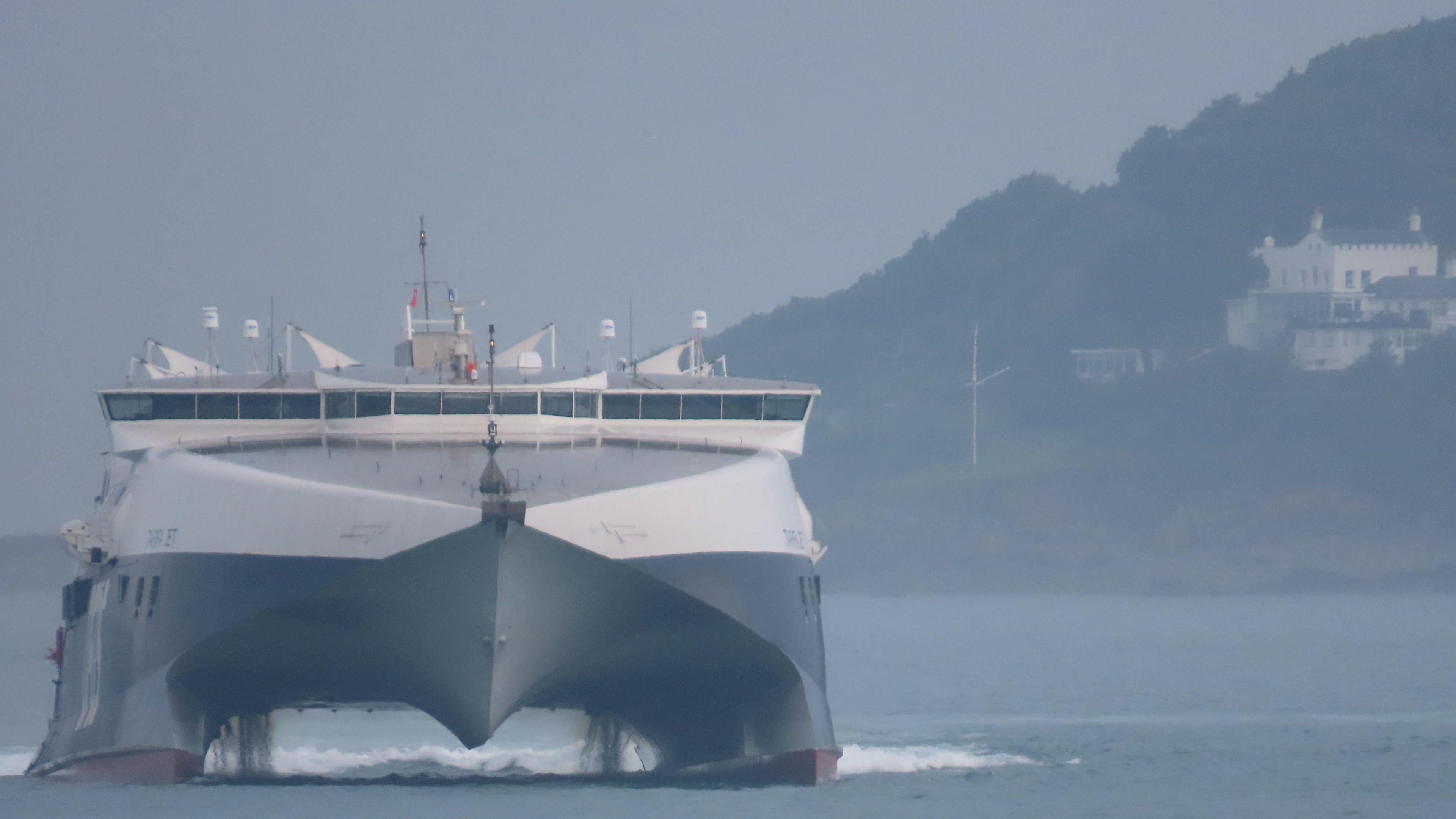 The front view of a DFDS ferry. To the right is land with a house on it. The ferry is black at the bottom and white at the top. It is moving on the water.