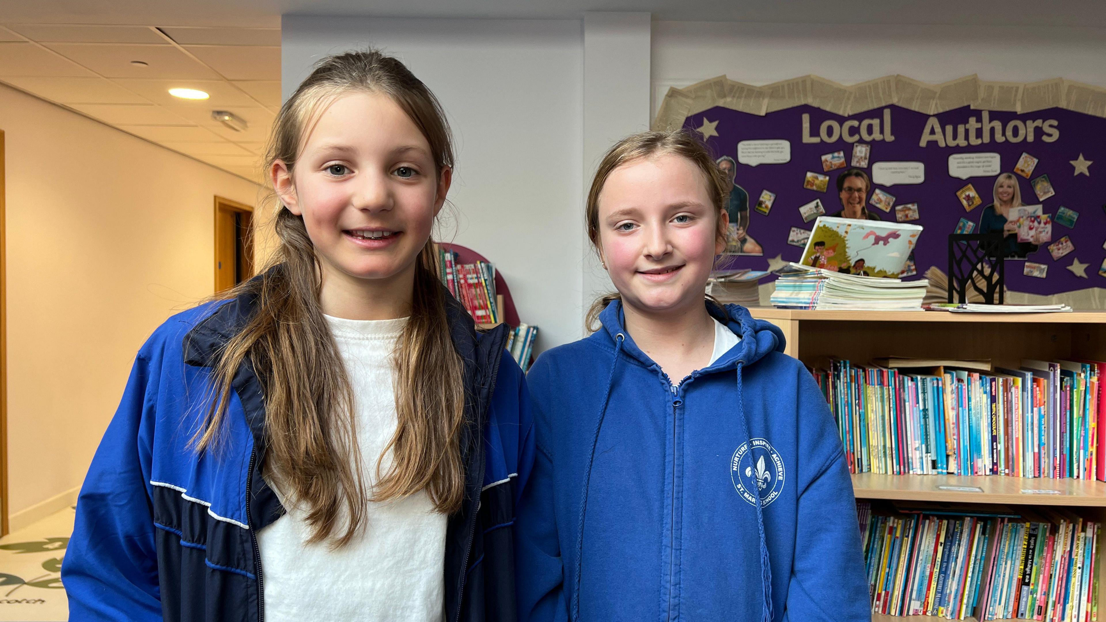 Annabella and Mia smile at the camera. Annabella has a blue jacket on which is unzipped and has long brown hair which comes over her shoulders and down to her chest. Mia is wearing a blue hoodie and has light blonde hair which is tied into a ponytail.