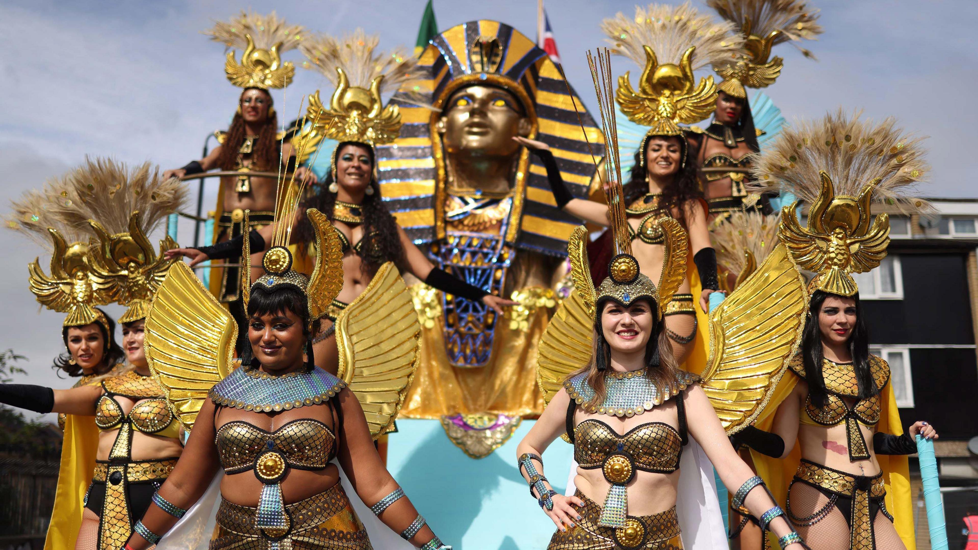 Parade of ancient Egyptian dancers wearing gold bikini tops and golden wings. Some of the women also have golden headdresses with feathers attached. There is also a Tutenkhamun death mask in the background. 