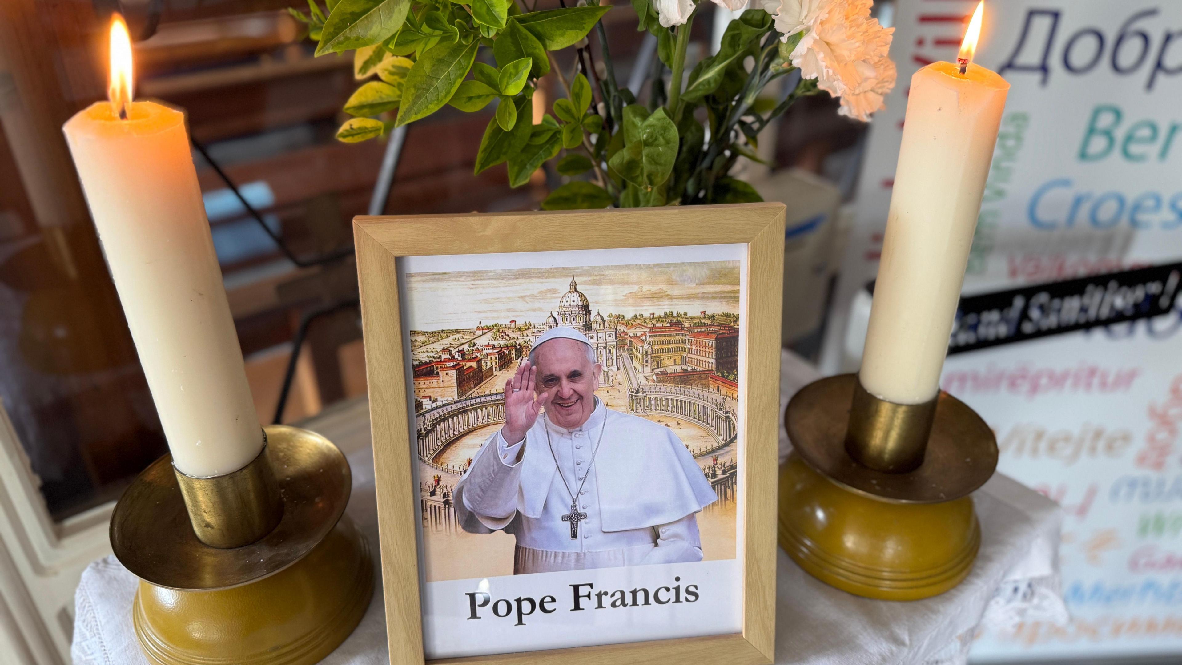 A picture of Pope Francis in a wooden frame, positioned on a table in the middle of two lit candles.
