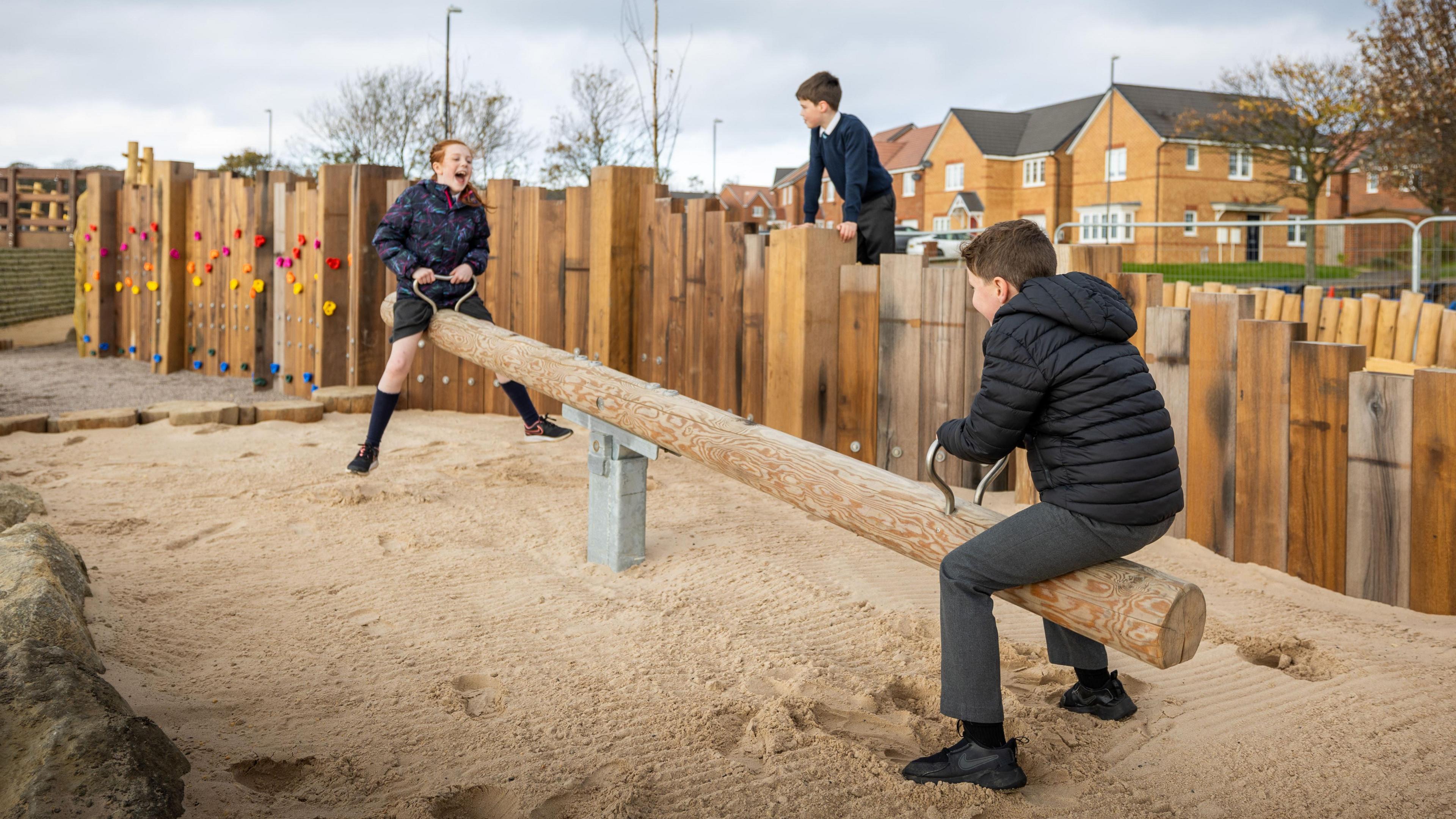 Children play on a wooden seesaw in the new park. Residential houses can be seen in the background.