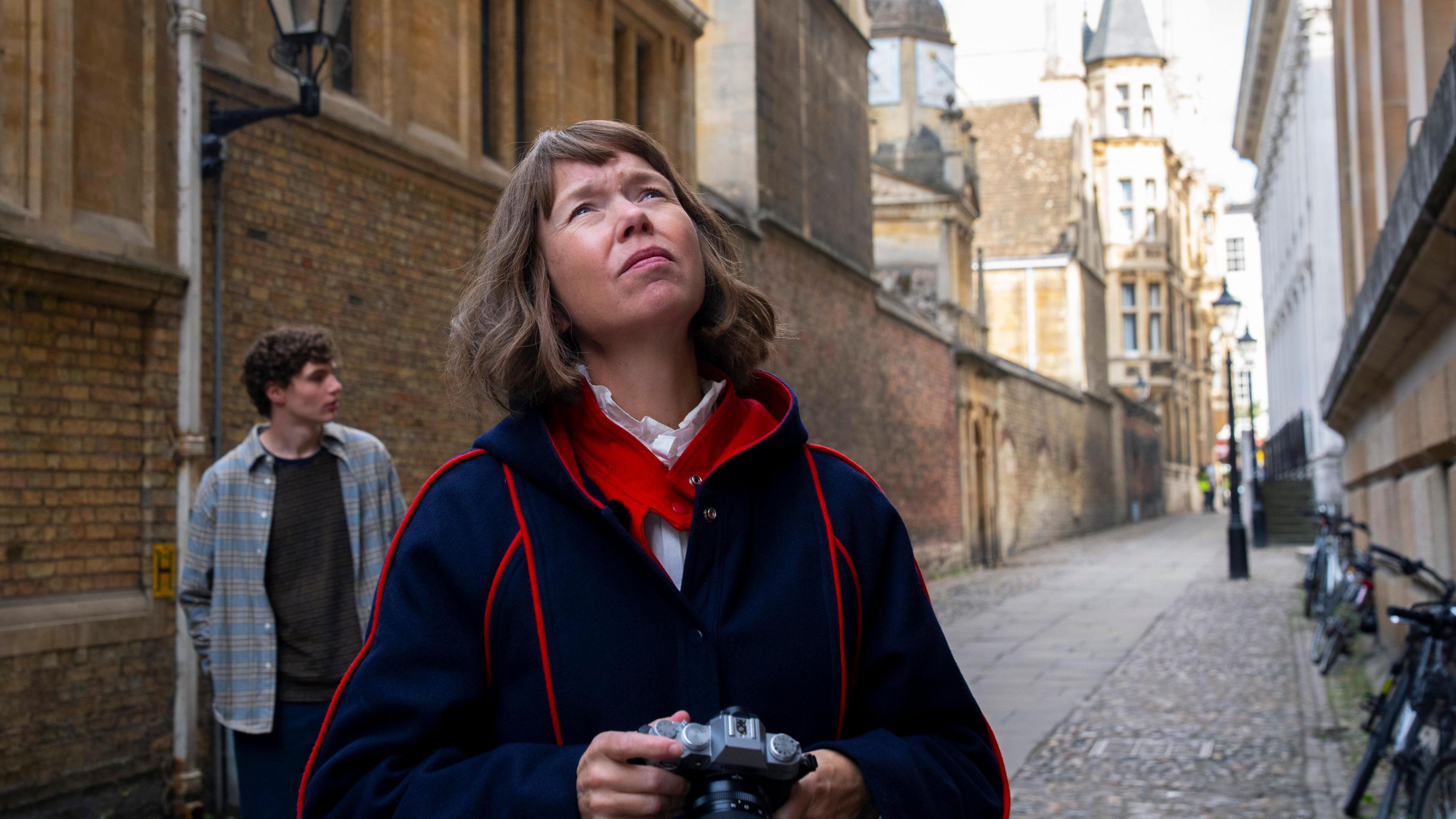 A woman in a blue coat with red lining stands in an alleyway in Cambridge, behind her on her right is a young man with brown hair wearing a plaid shirt.