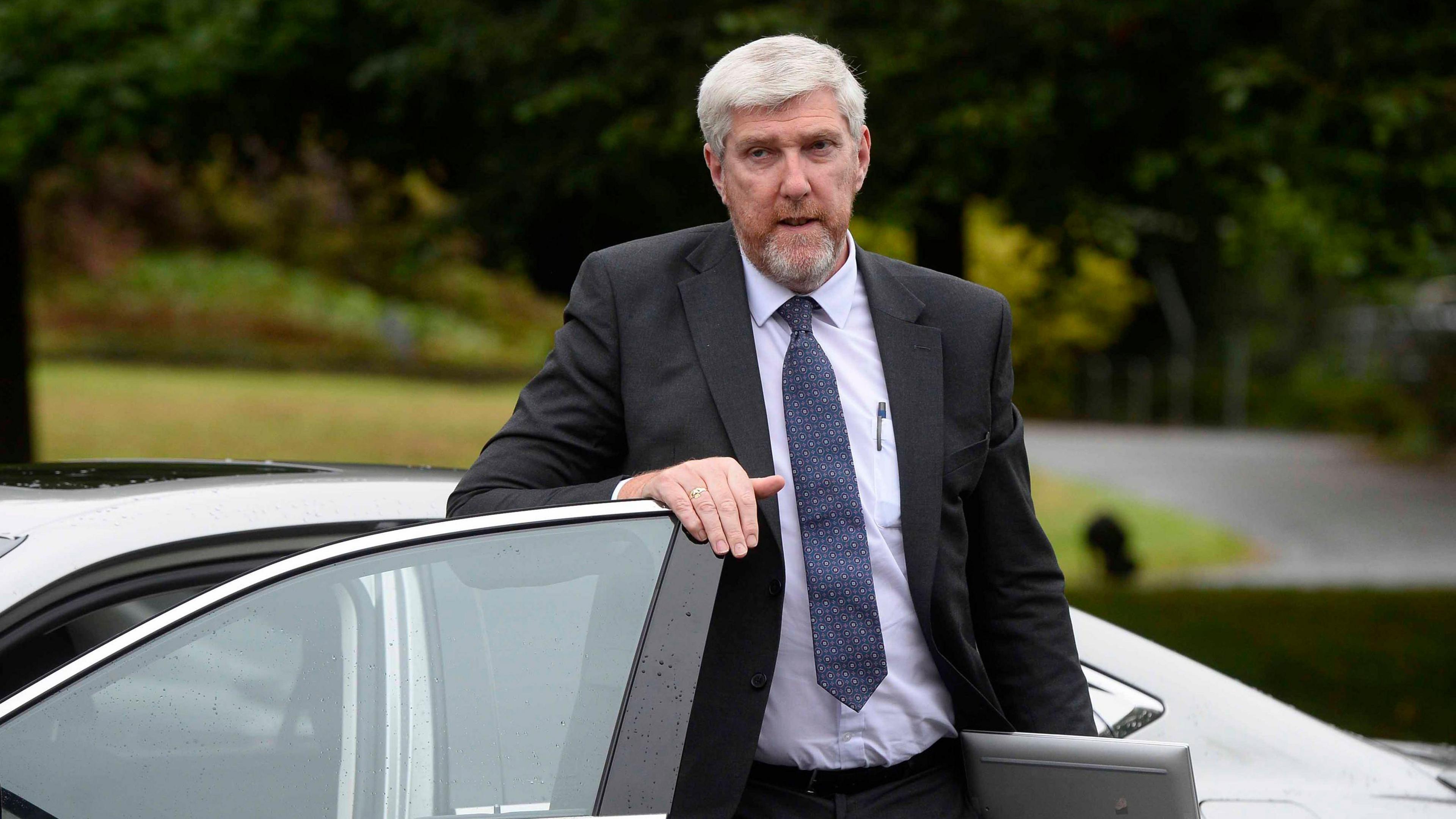 Infrastructure minister, John O'Dowd, getting out of a car wearing a black suit with a white shirt and blue tie. He is carrying a laptop.