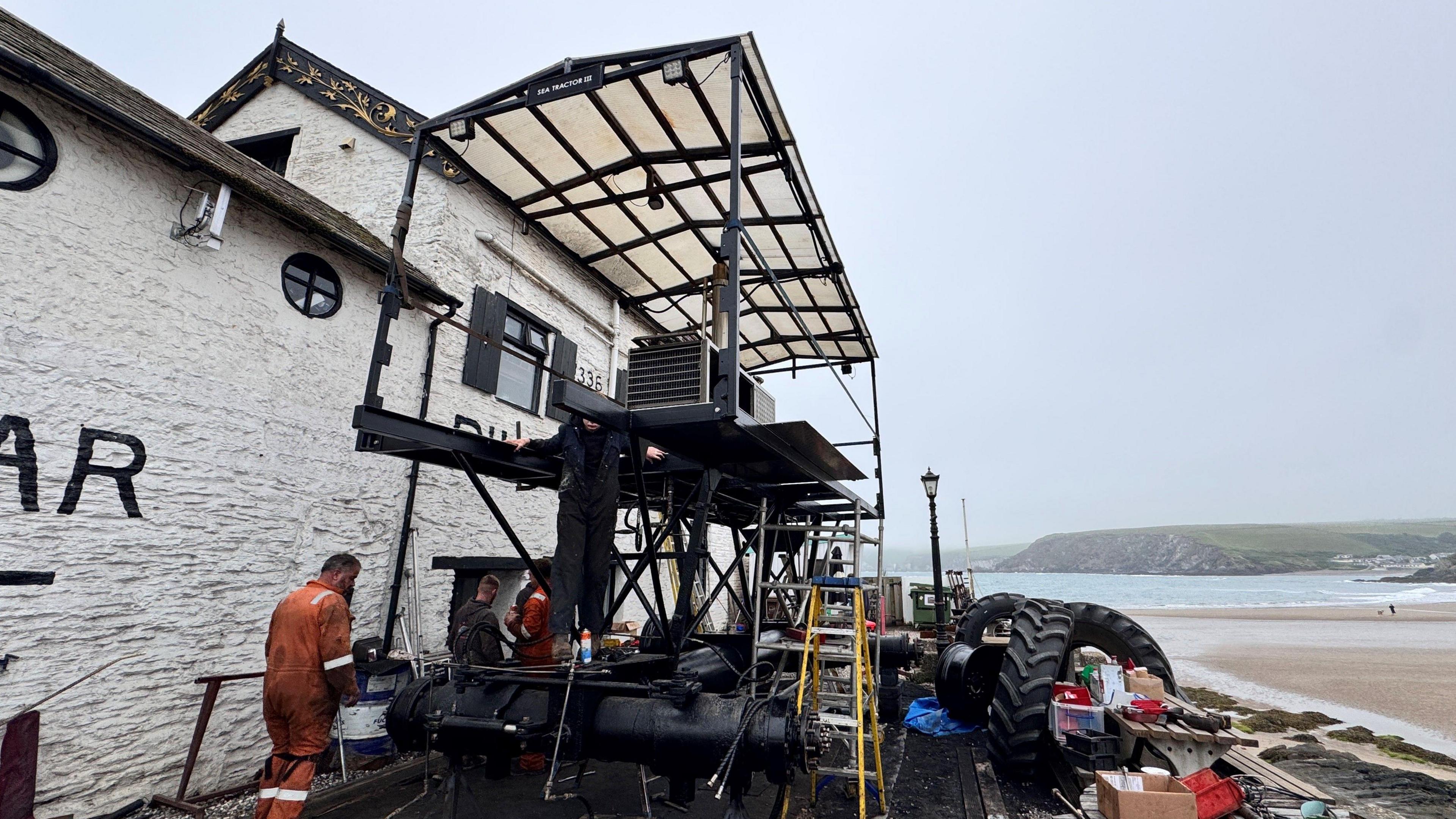 The picture shows a group of men working on a sea tractor just off the beach. They are doing restoration work on the vehicle which is painted black and had four agricultural style wheels. There is a pub next to it and the sea in the background.