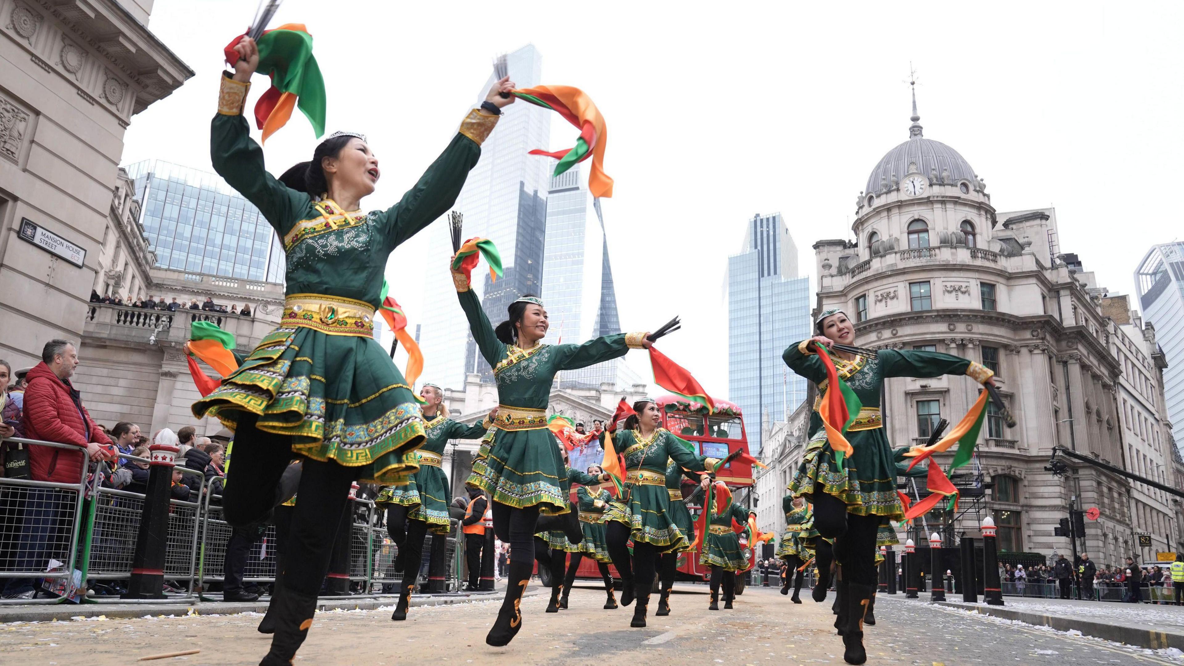 A group of dancers in vibrant green and gold costumes perform during the Lord Mayor's Show, waving colourful orange and green scarves. They dance energetically down a London street lined with historic buildings and modern skyscrapers, while spectators watch from the pavements.