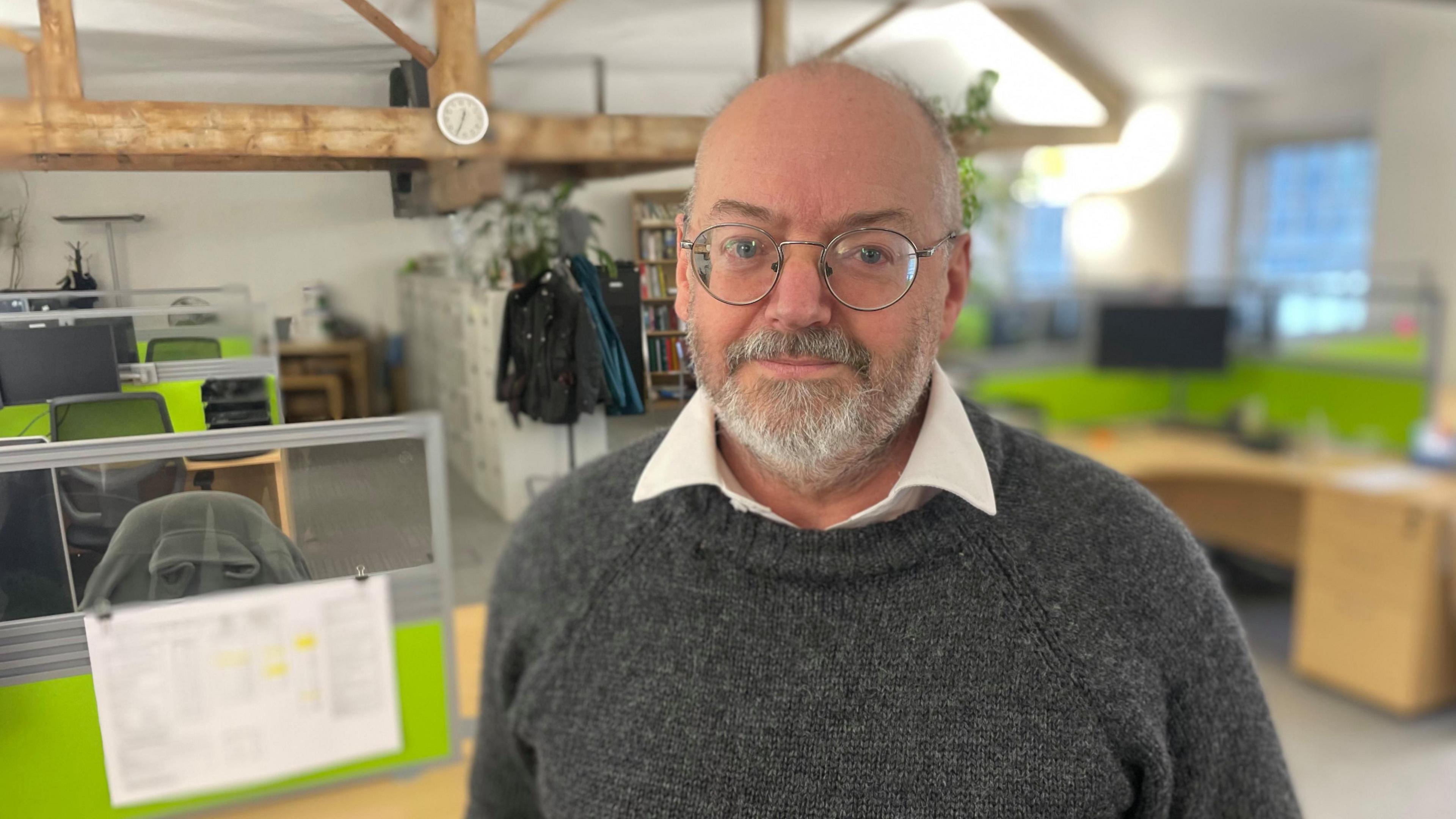 Simon Edwards (round glasses, beard, grey jumper, cream shirt) stands in the offices of Merebrook Consulting, which has wooden beamed ceilings and modern office furnishings behind.