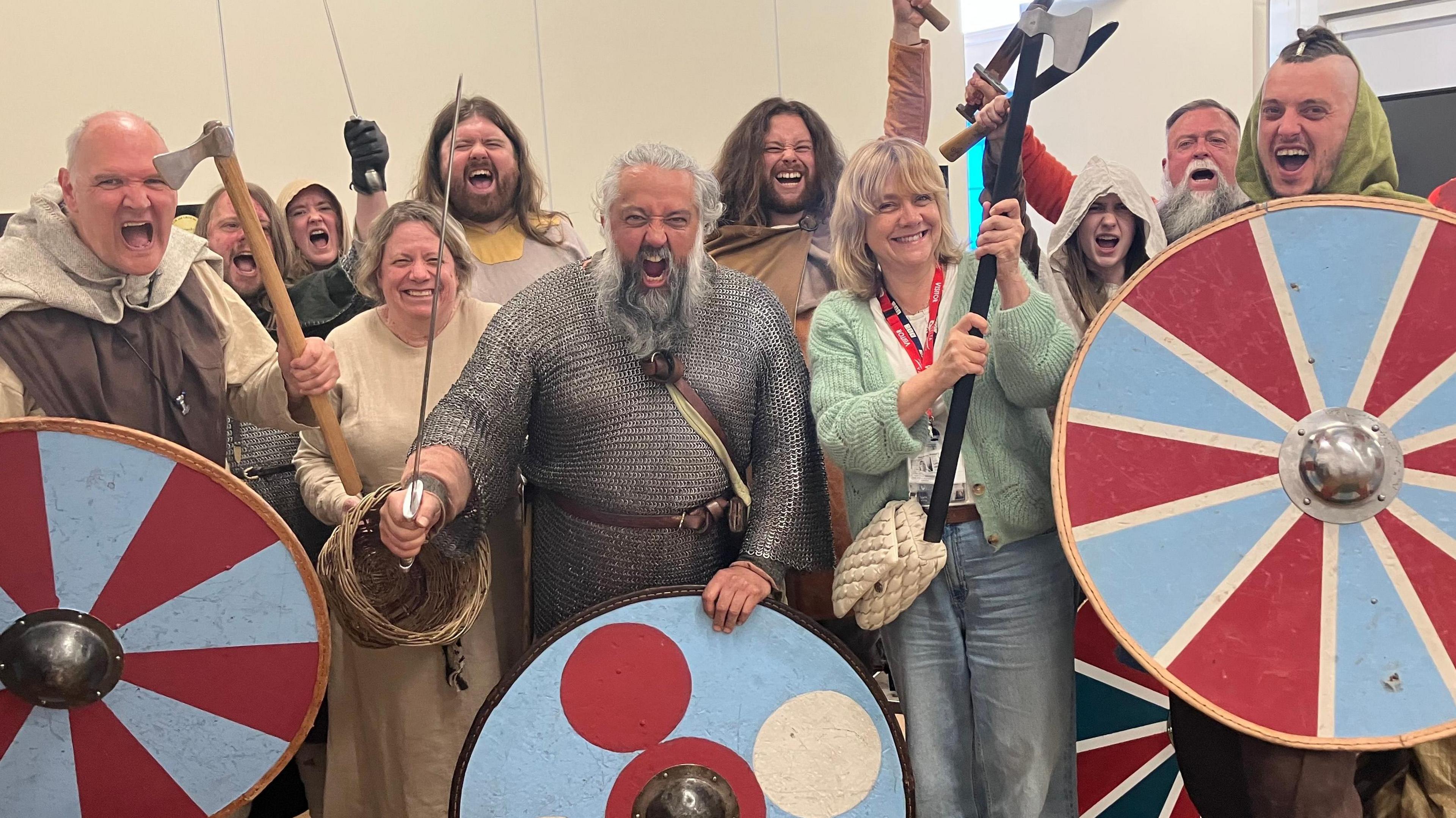 A group of 11 men and women dressed as Vikings, including a man in chain-mail armour and some holding shields and swords, cheer as they stand in a tent.