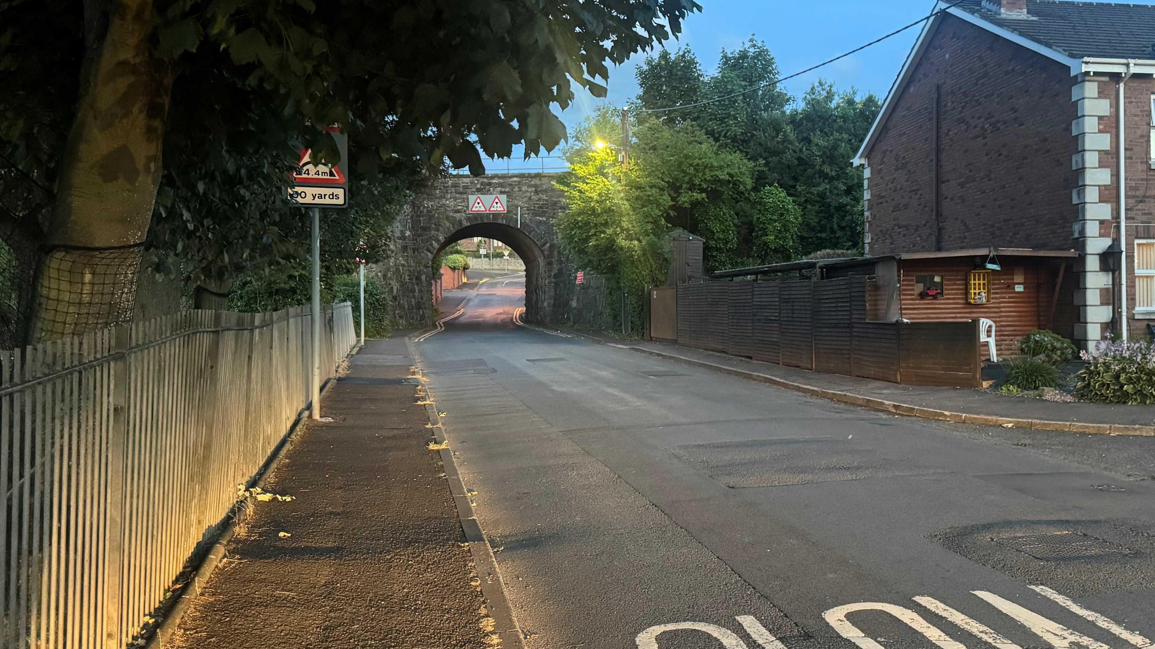 A narrow, empty road leads to an opening underneath a bridge. 'SLOW' is painted in capital letters on the road. There is a residential home on the right of the road and a metal fence on the left.