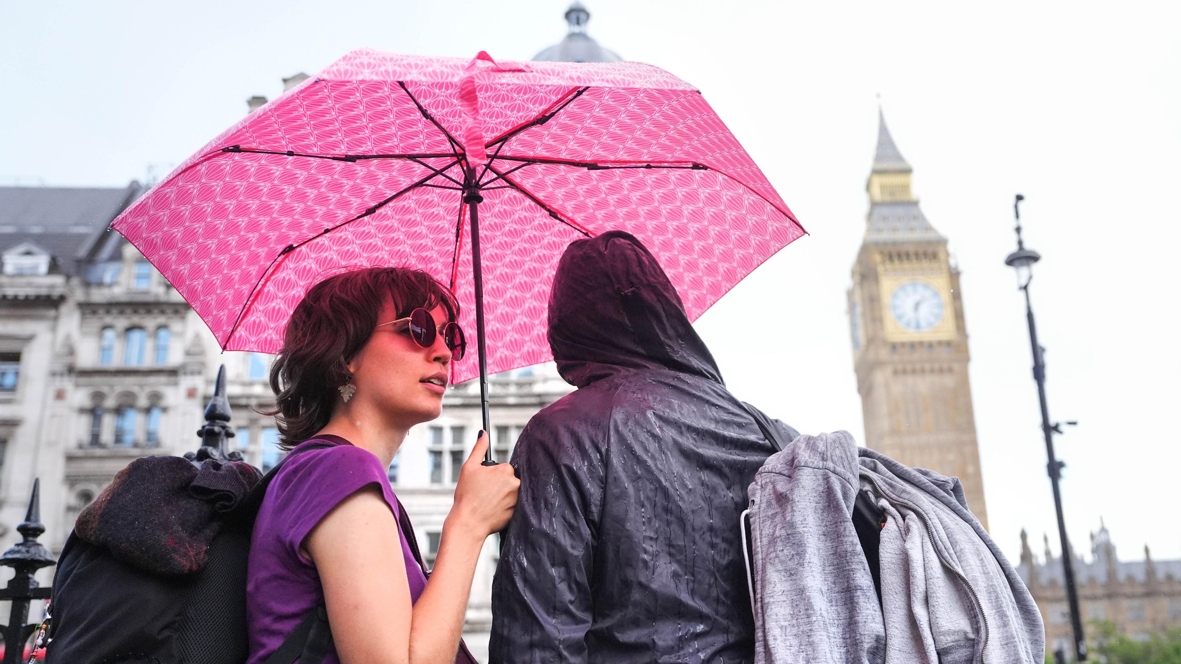 Two people stand under a pink umbrella sheltering from the rain with the Westminster skyline in the background