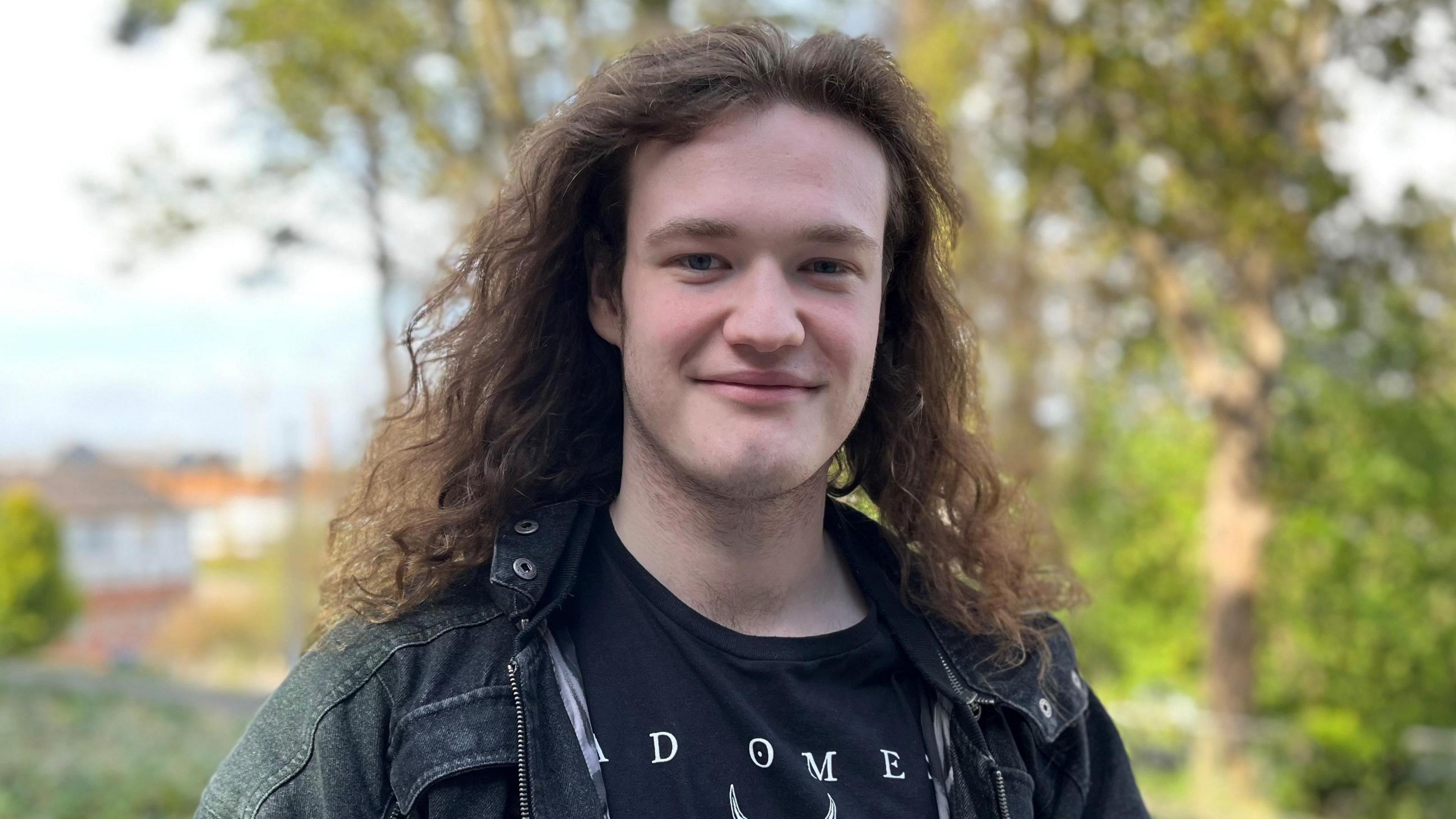 Liam is outside and looking at the camera. There are trees in the background. He has long hair and wears a dark denim jacket and black t-shirt