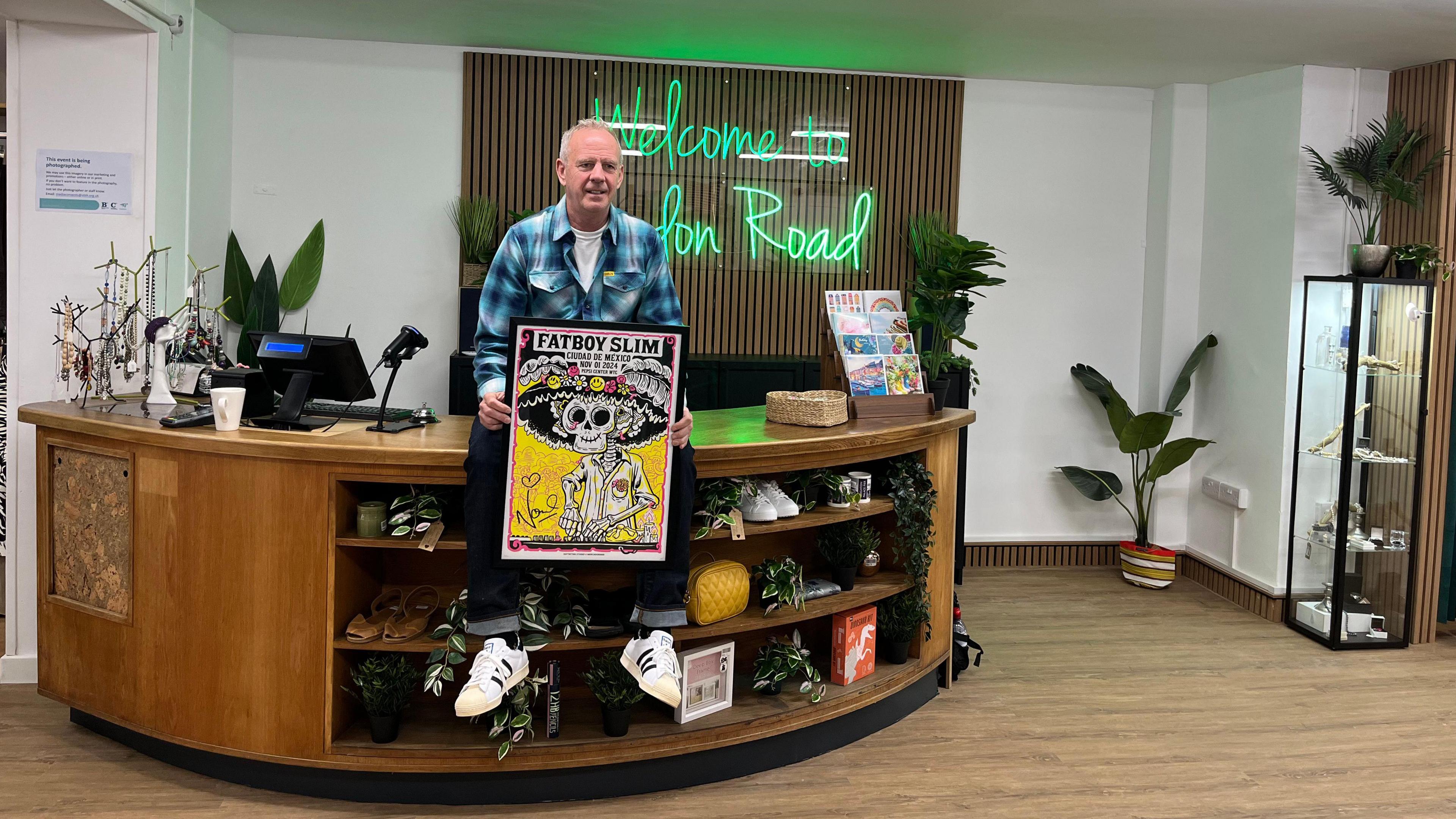 Fatboy Slim sitting on the counter of the London Road Martlets hospice charity shop holding a poster of his artwork