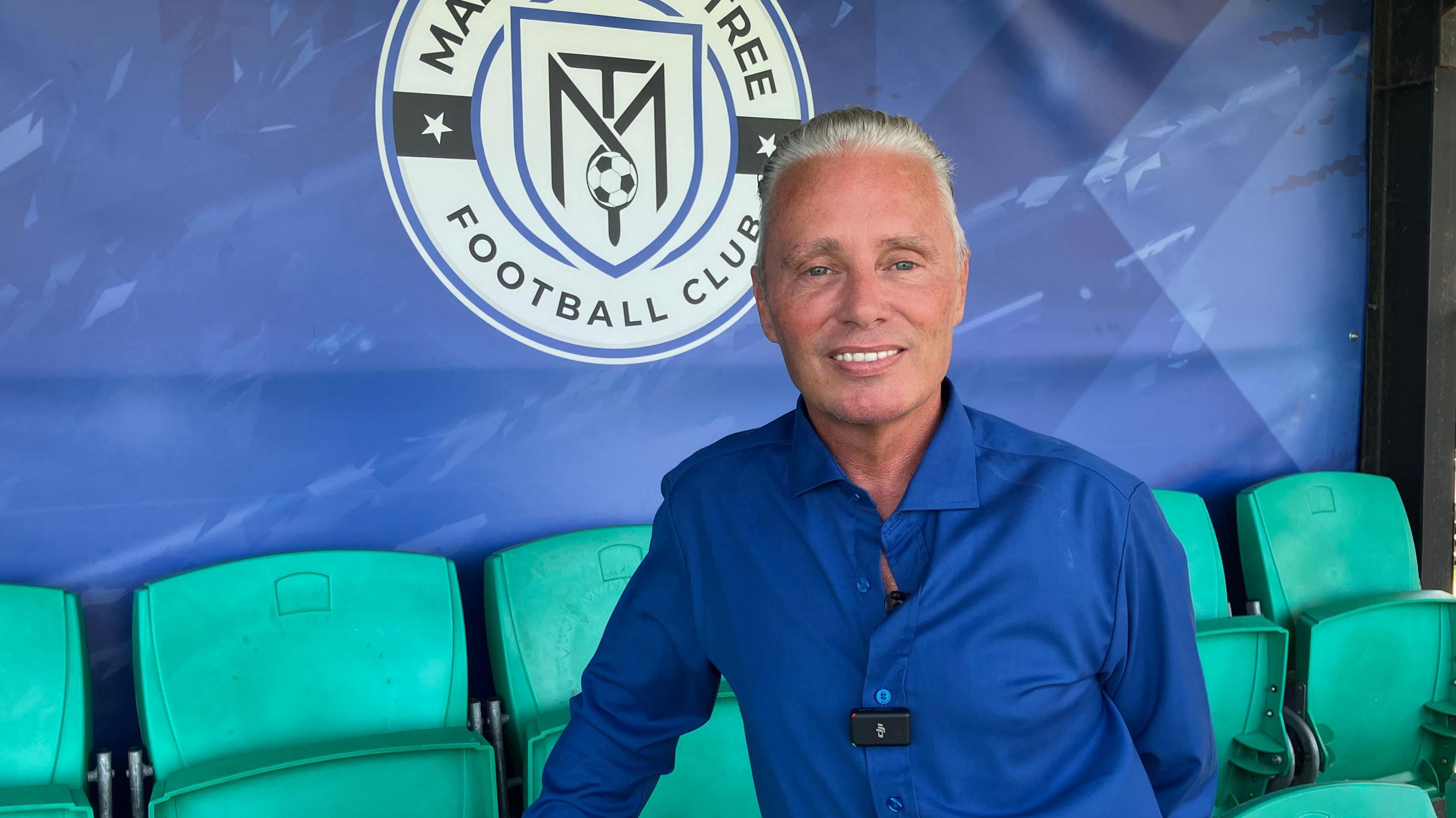 A smiling Barrie Drewitt-Barlow looking straight at the camera. He is sitting on a green seat in a football stadium in front of a blue wall with the logo of Maldon & Tiptree Football Club on it.