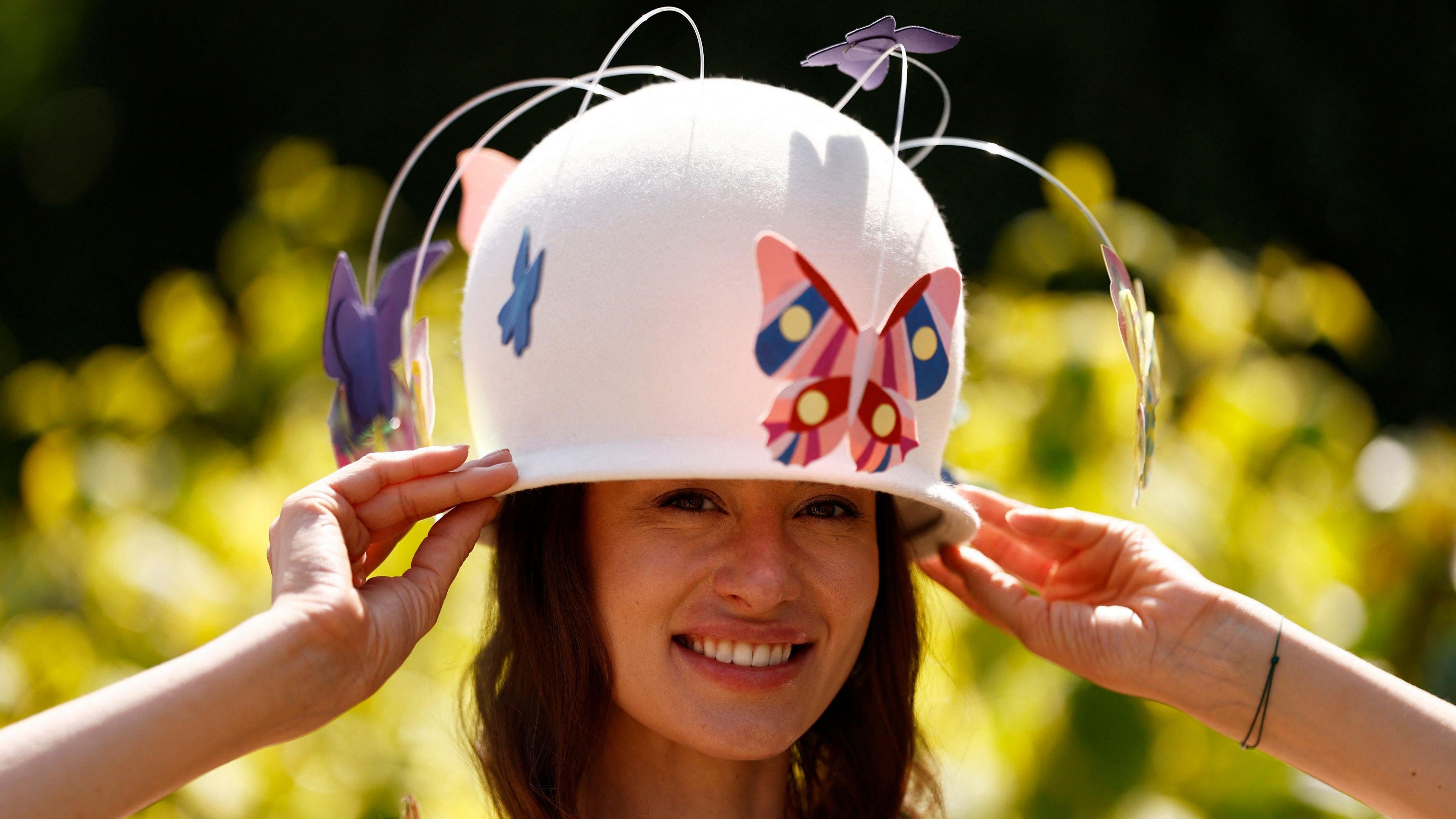 A woman wearing a white hat which is a play on a horse racing helmet, with butterflies hanging below it.
