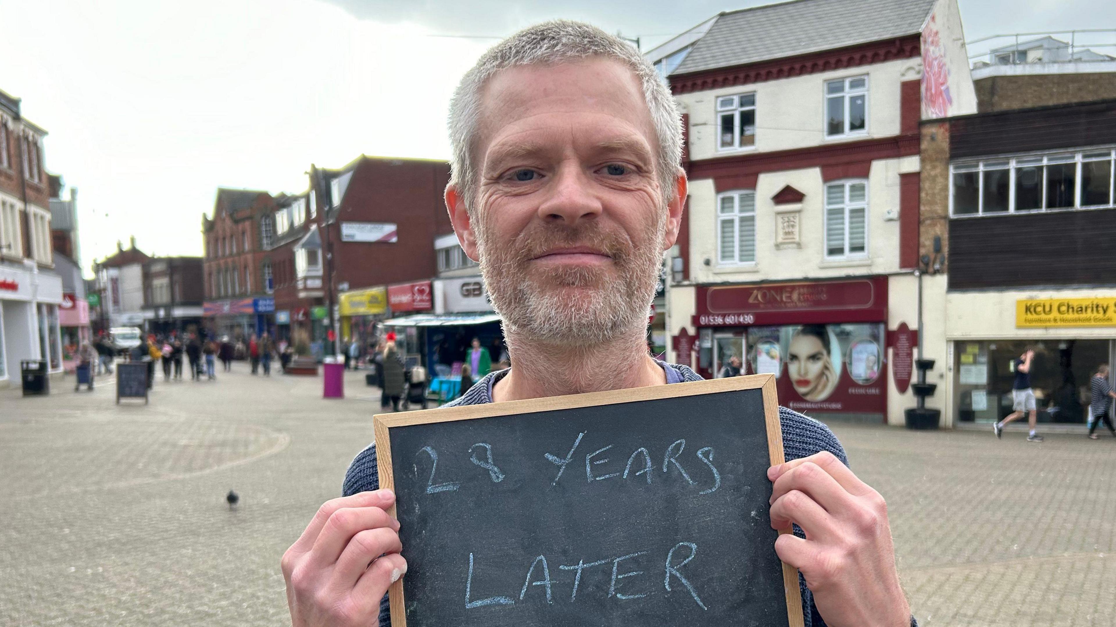 A man holding up a board which reads "28 Years Later". He is standing in Kettering town centre