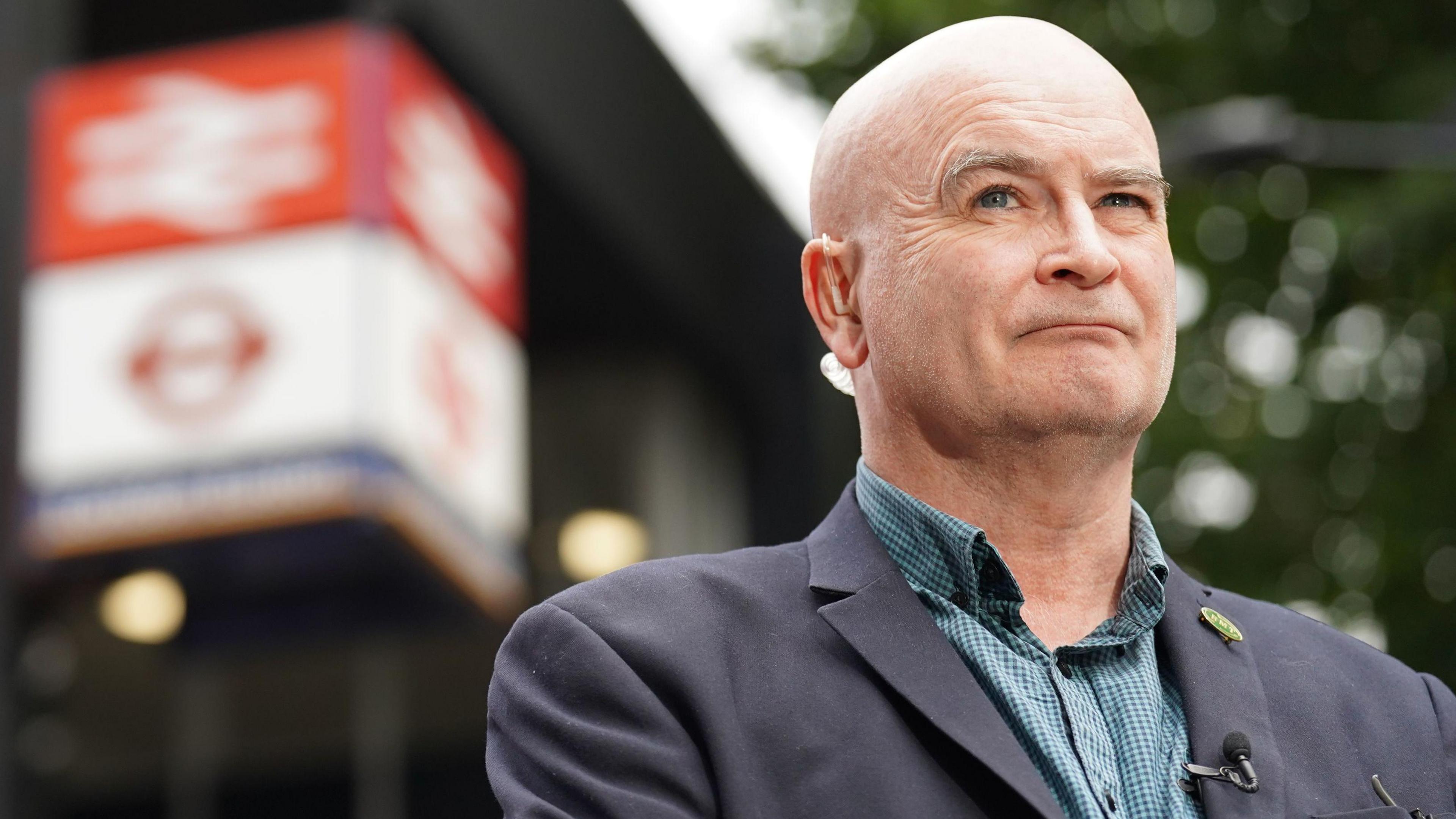 Mick Lynch stands waiting for an interview on the picket line outside London Euston train station. There is train station sign behind him and he is wearing a suit and has a visible earpiece