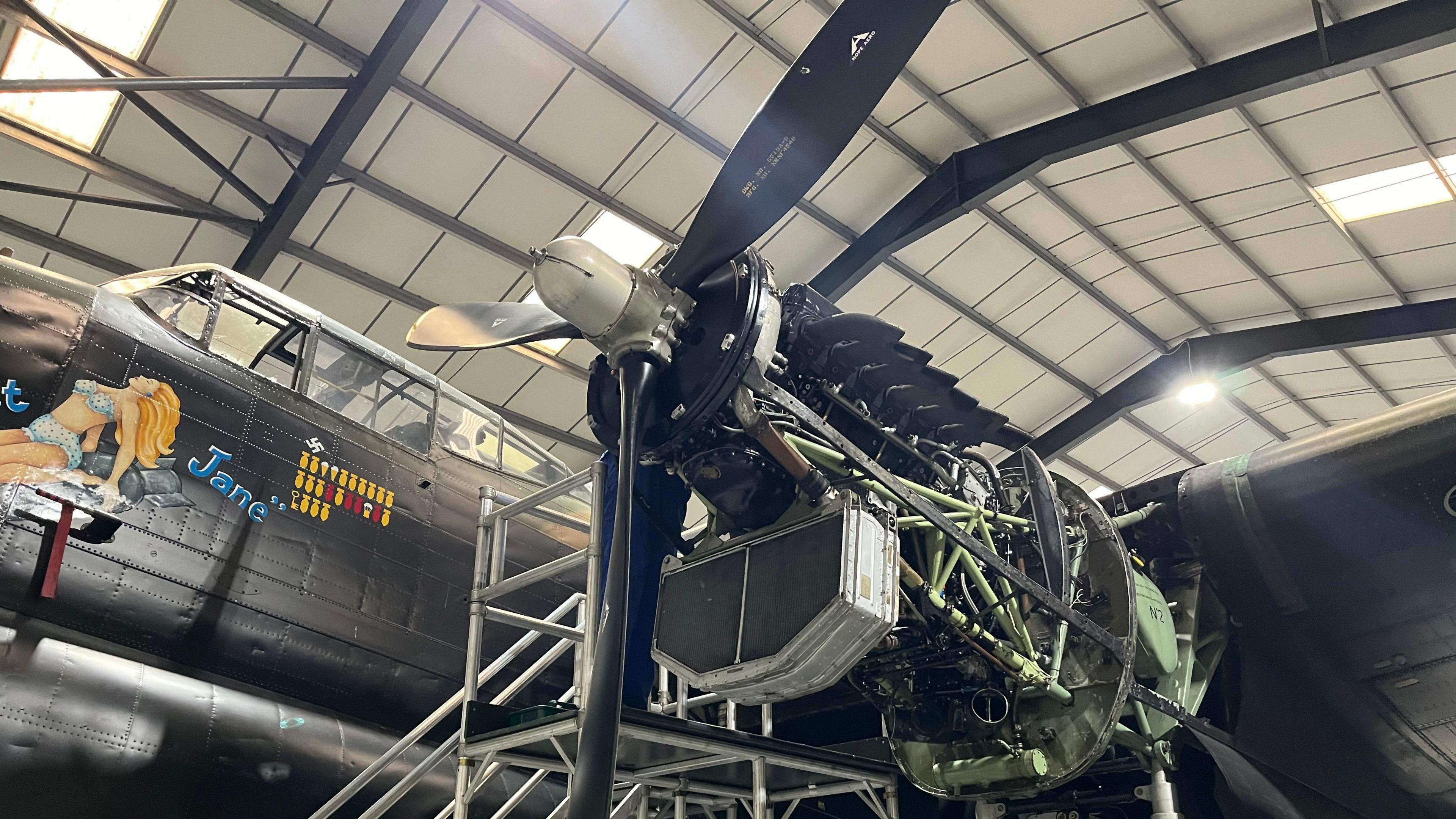 A side view of Just Jane in a warehouse. The aircraft's engine is exposed, revealing mechanical parts, pipes and wiring. 