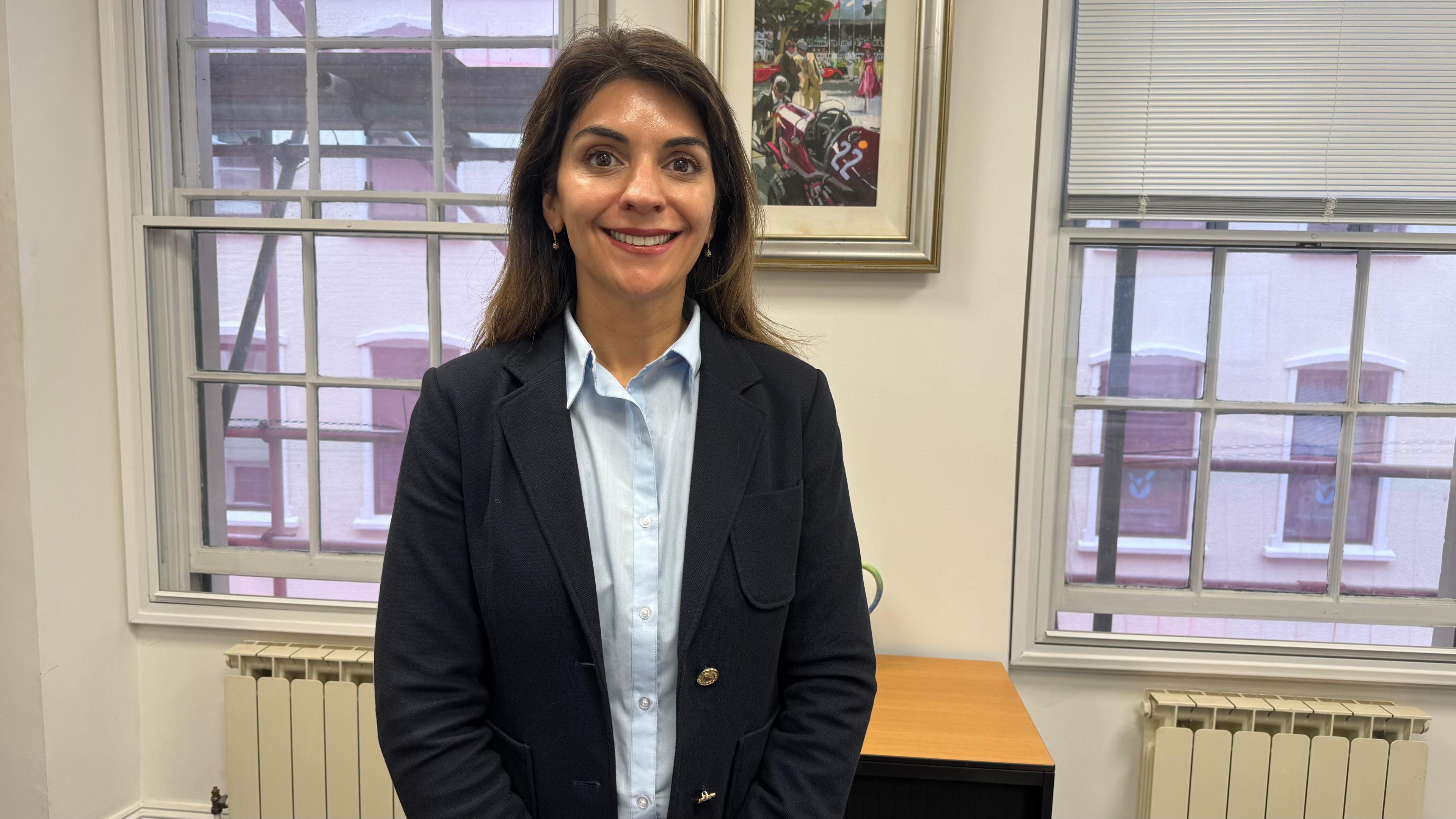 Advocate Natasha Newell. Partner at No.10 Advocates wearing a blue shirt and blazer. She is standing in her office in front of a window.