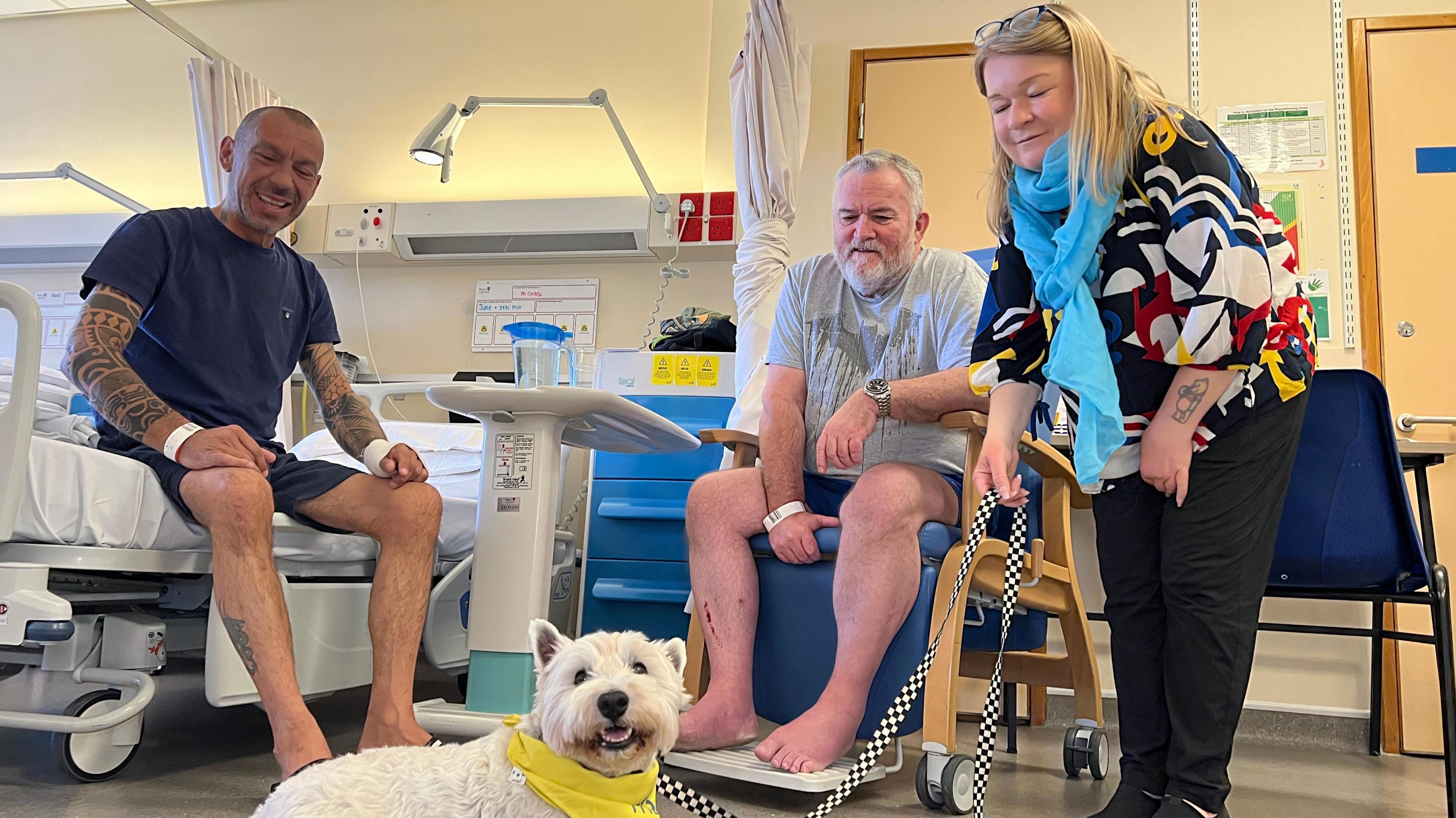 Frankie lies down in front of two patients, one man sets on a hospital bed and another is in a chair as Anna leans over holding Frankie's lead