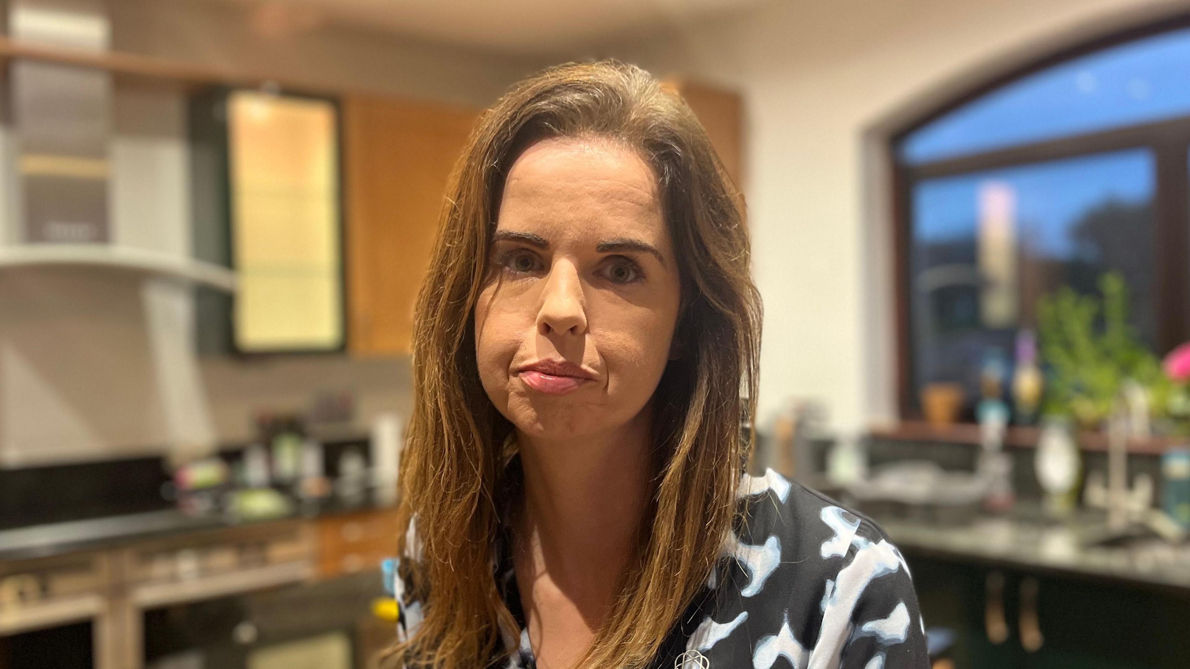 Denise Heavey is sat in her kitchen, looking at the camera. She is wearing a black and white shirt.