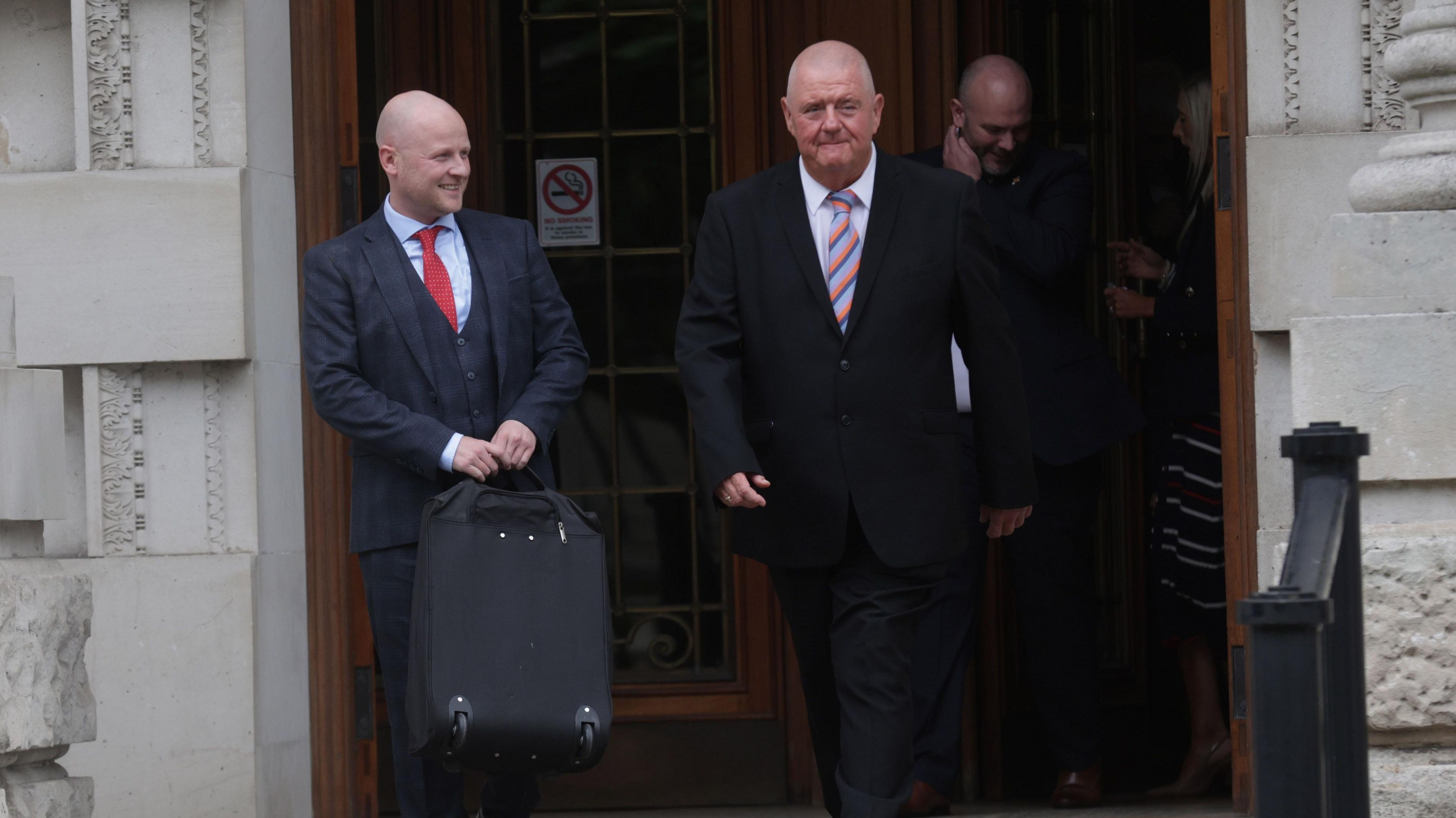 Jamie Bryson leaves court. He is bald, carrying a suitcase, wearing a dark suit, light blue shirt and red tie. A man is beside him, he is bald, wearing a dark suit, white shirt and multi-coloured tie.
