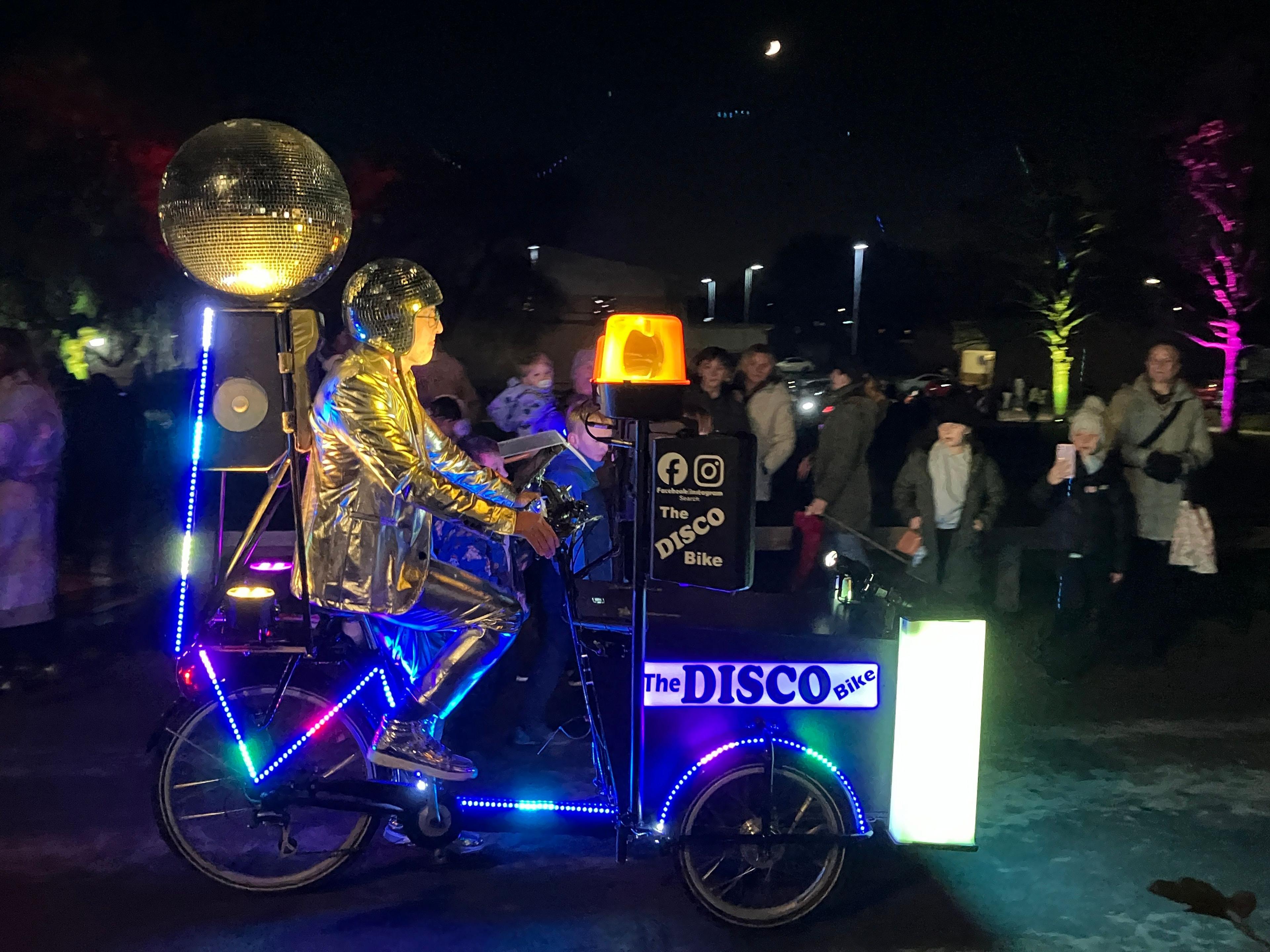 A man riding a brightly lit Disco Bike, with a gold glitter ball on the back.