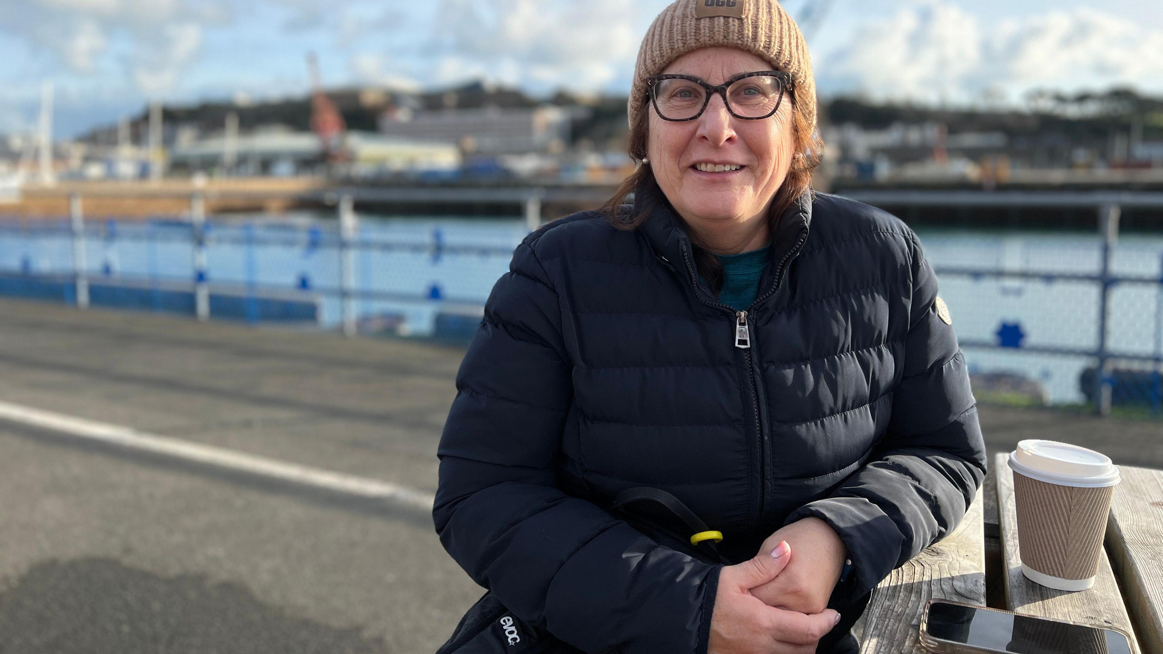 A woman in a black puffer jacket with shoulder-length brown hair, black rimmed glasses and a light brown beanie hat sits with a coffee at a wooden table, with the docks behind her.