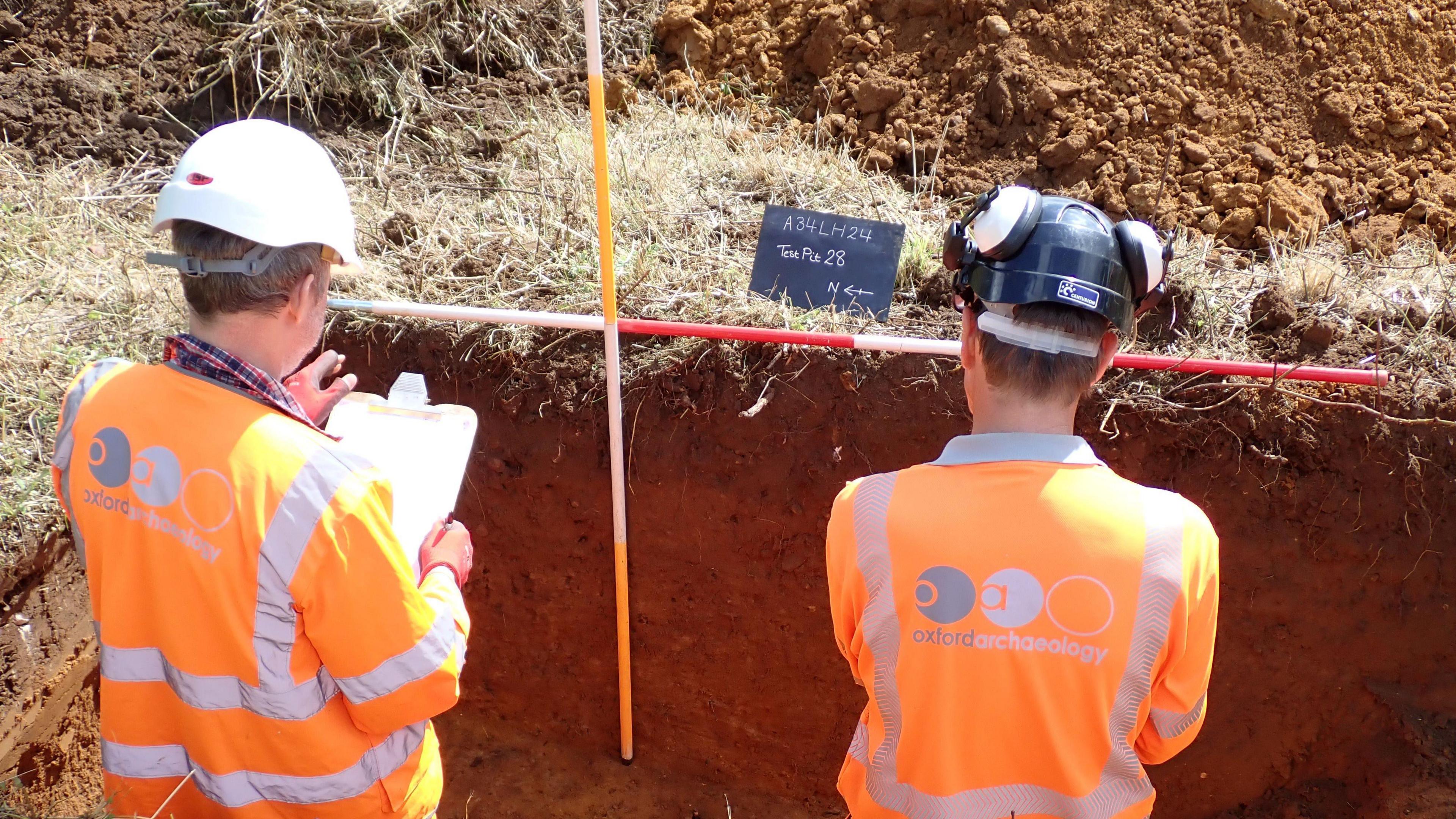 Two men in high-vis tops and hard hats standing in a trench. One of them is holding a clipboard