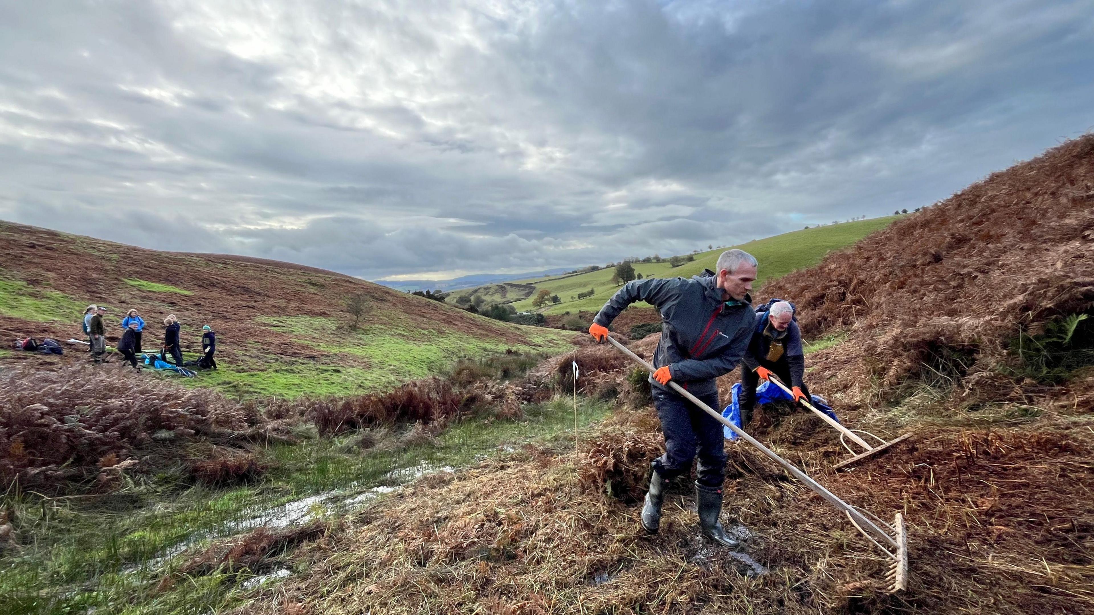 Two volunteers are in the foreground, using rakes on brown grass and earth. In the background are five other people, standing on grass on the side of a hill. Further in the distance are rolling hills with trees and bushes